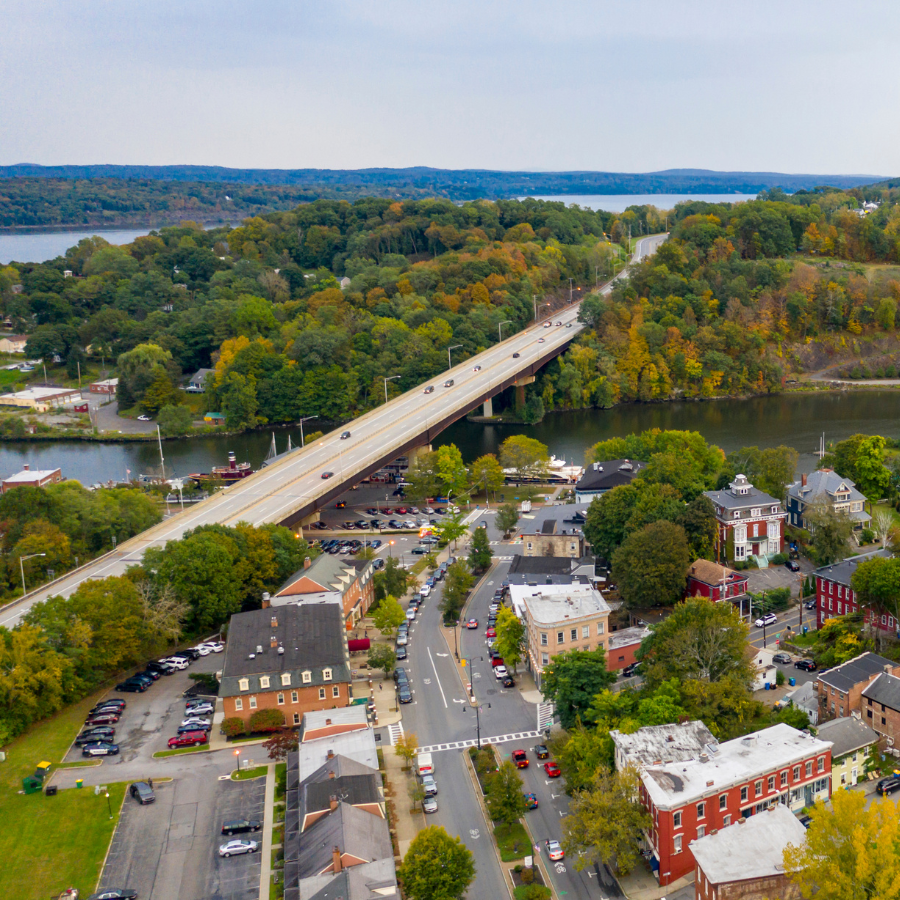Aerial view of a town with a bridge over a river, tree-lined streets, and autumn-colored hills.