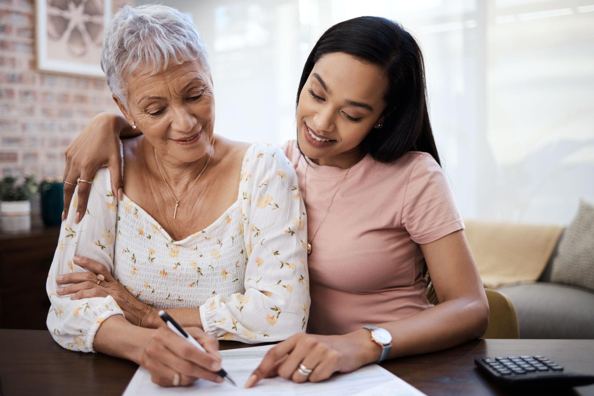 Two women reviewing documents at a table, one writing while the other leans in and smiles