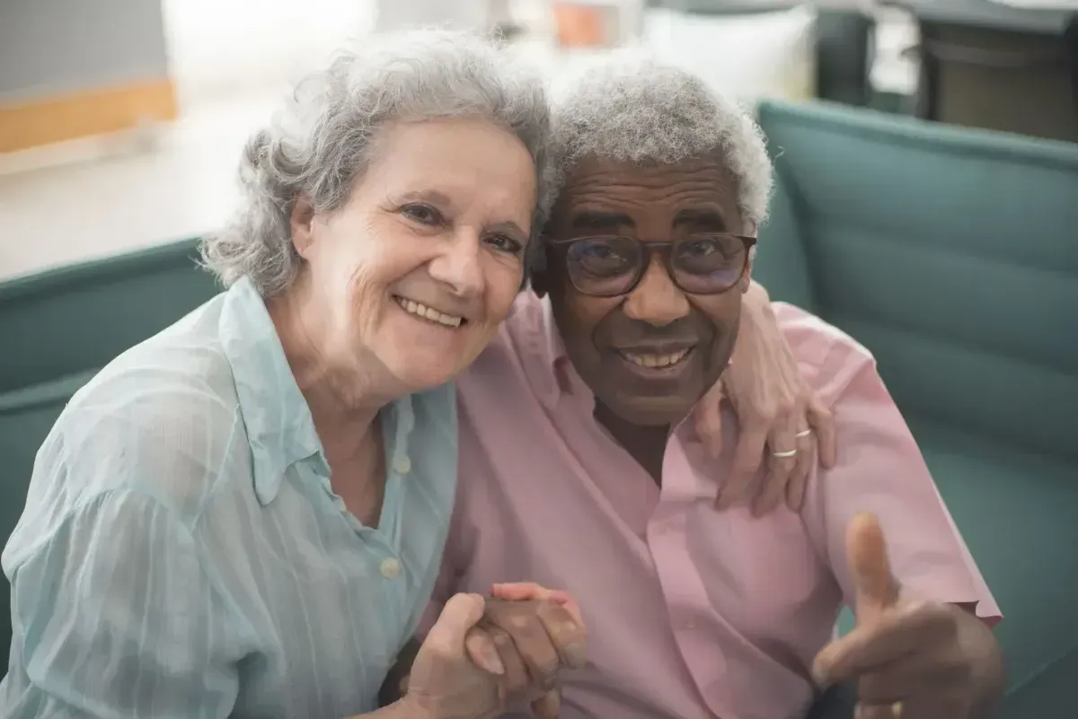 Two smiling people sit on a couch, hugging and giving a thumbs-up.