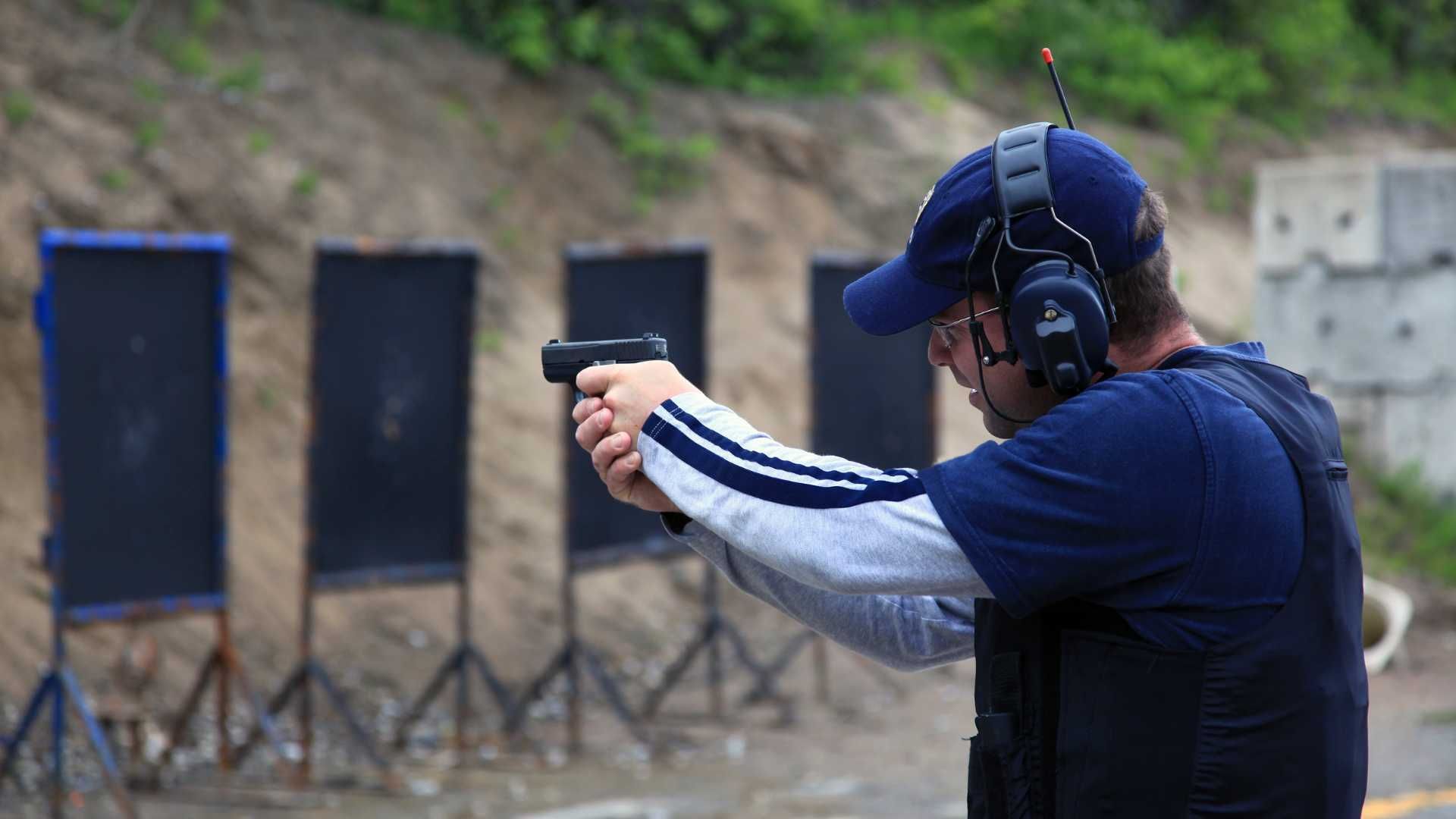 A person wearing a blue shirt, cap, and ear protection practices firing a handgun at a row of targets on a firing range.