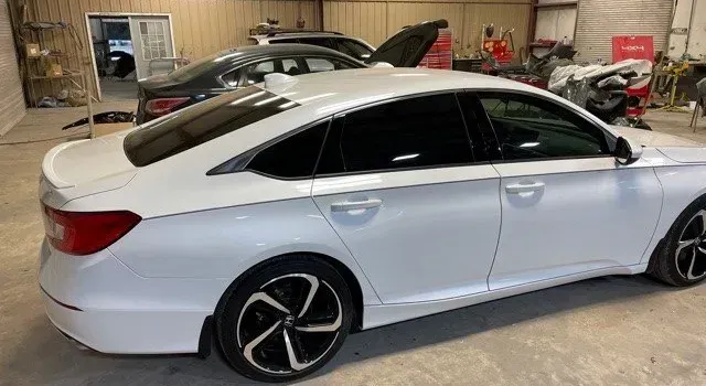 White sedan in a garage with its trunk open, parked beside motorcycles and other vehicles