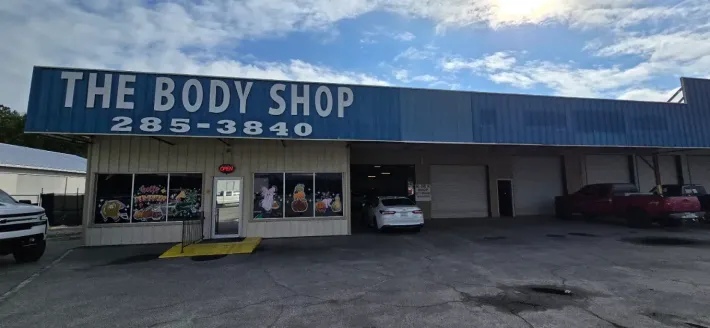 Storefront for “The Body Shop” with blue awning and parked cars in front.