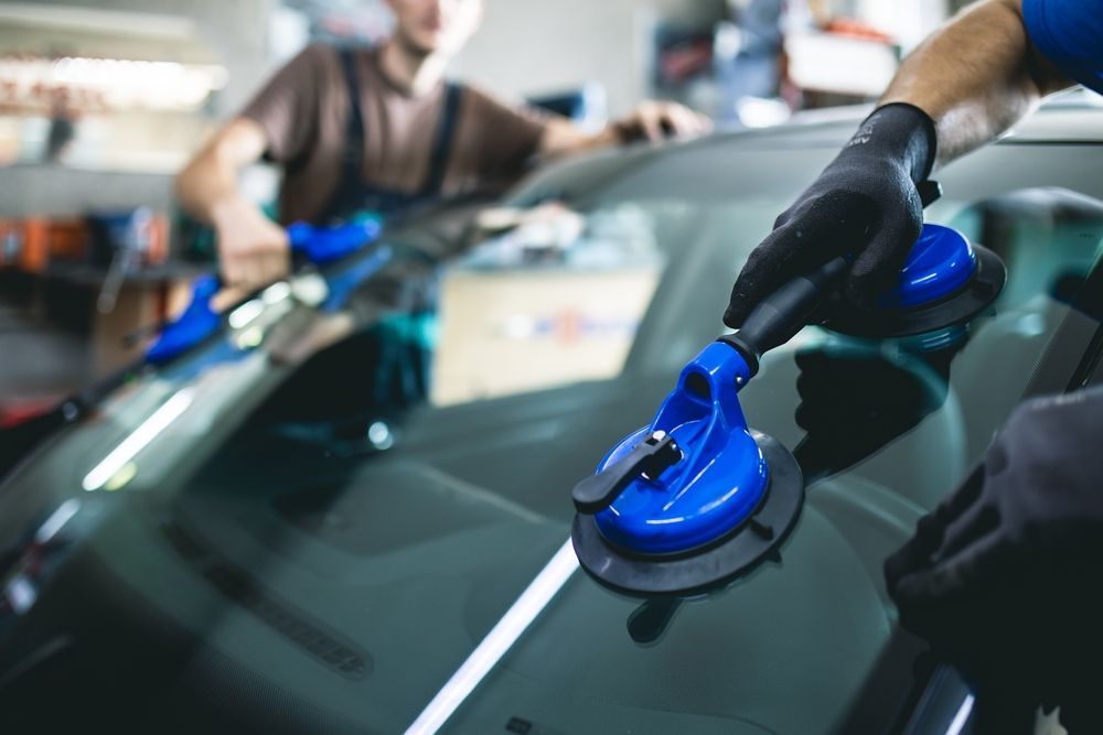 Gloved worker polishing a car windshield with a blue suction buffer in an auto shop