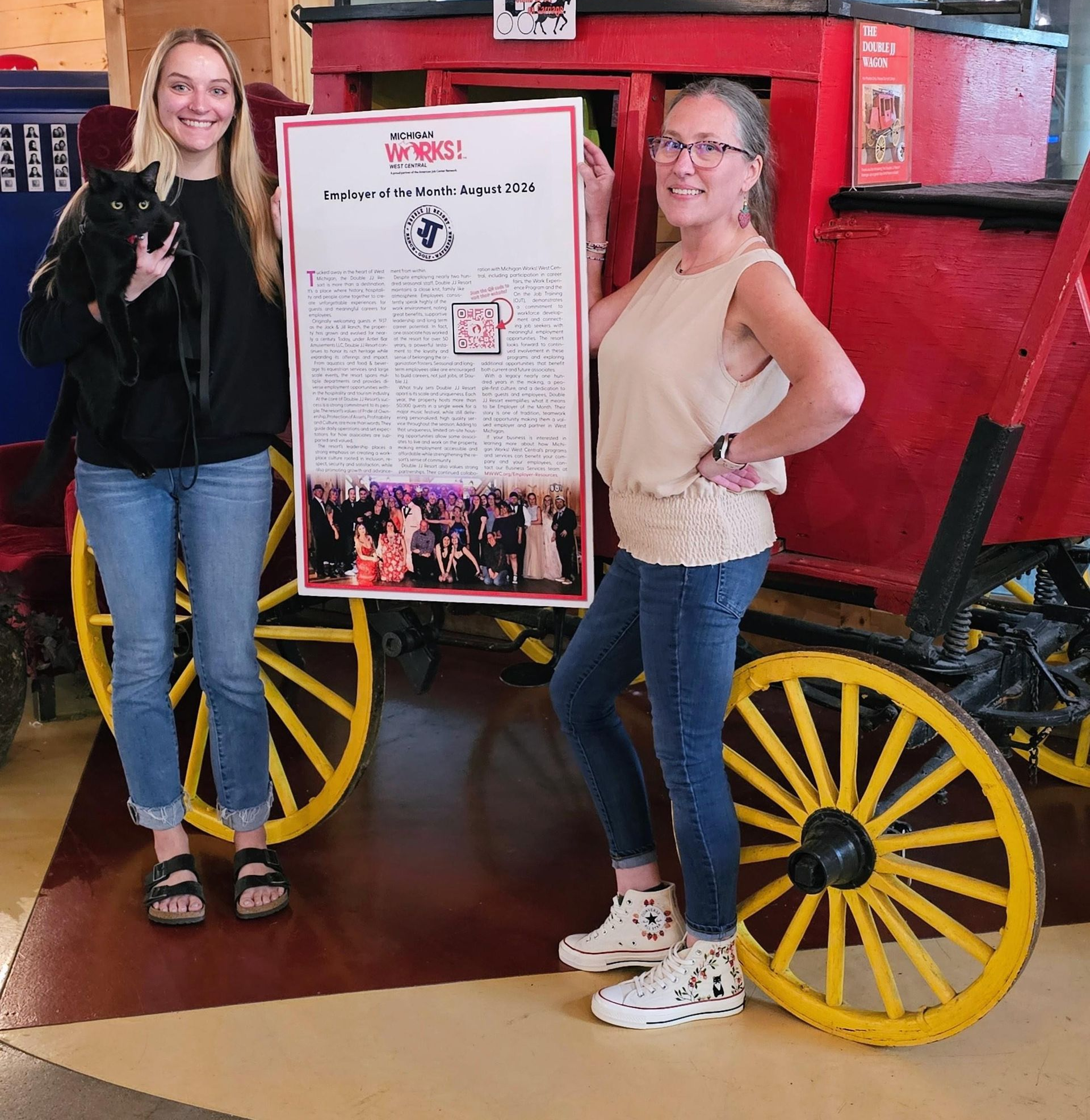 Two women standing beside a red wagon with yellow wheels and a poster display indoors