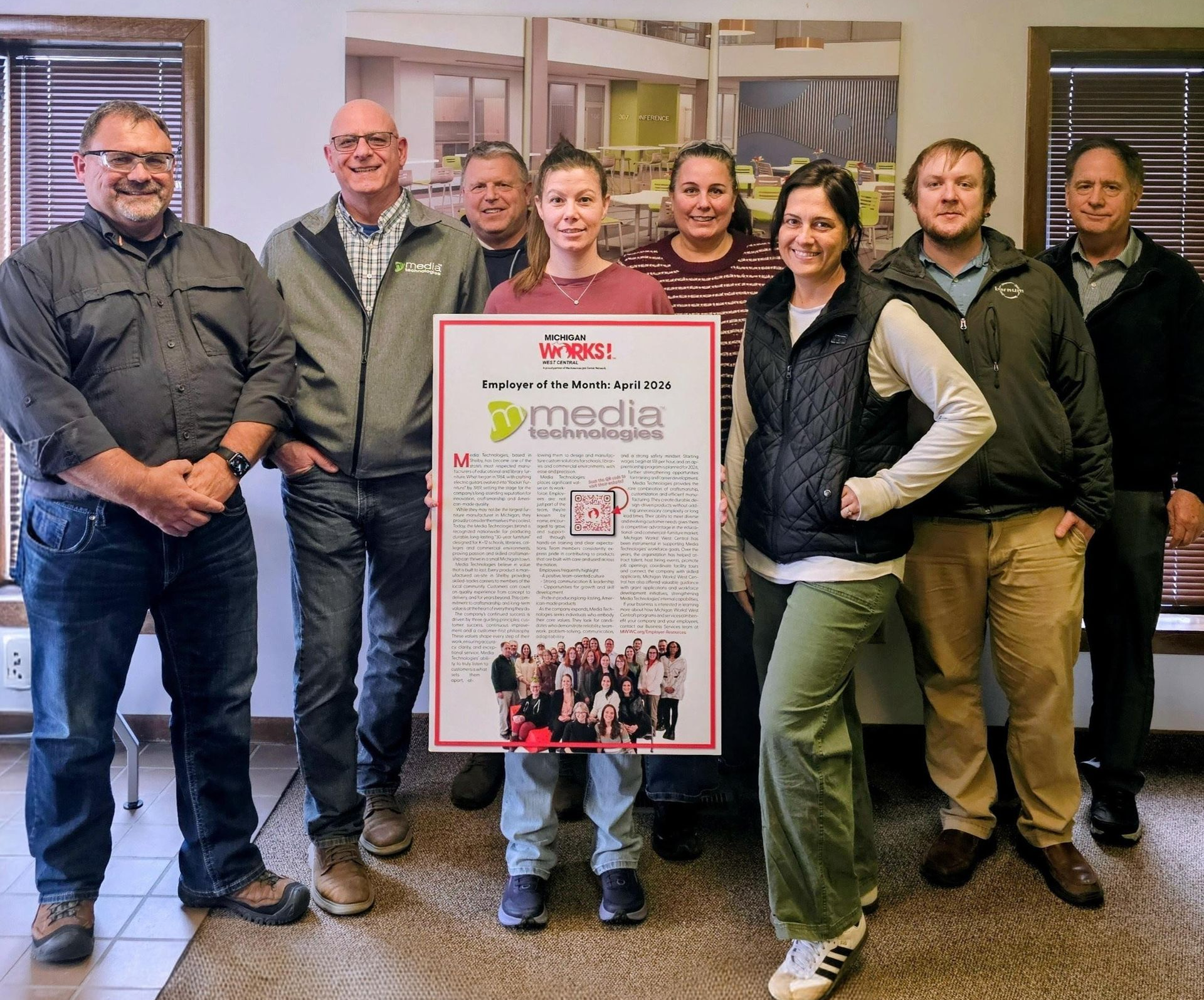 Group of people standing indoors, holding a poster with red border and a crowd photo at the bottom.