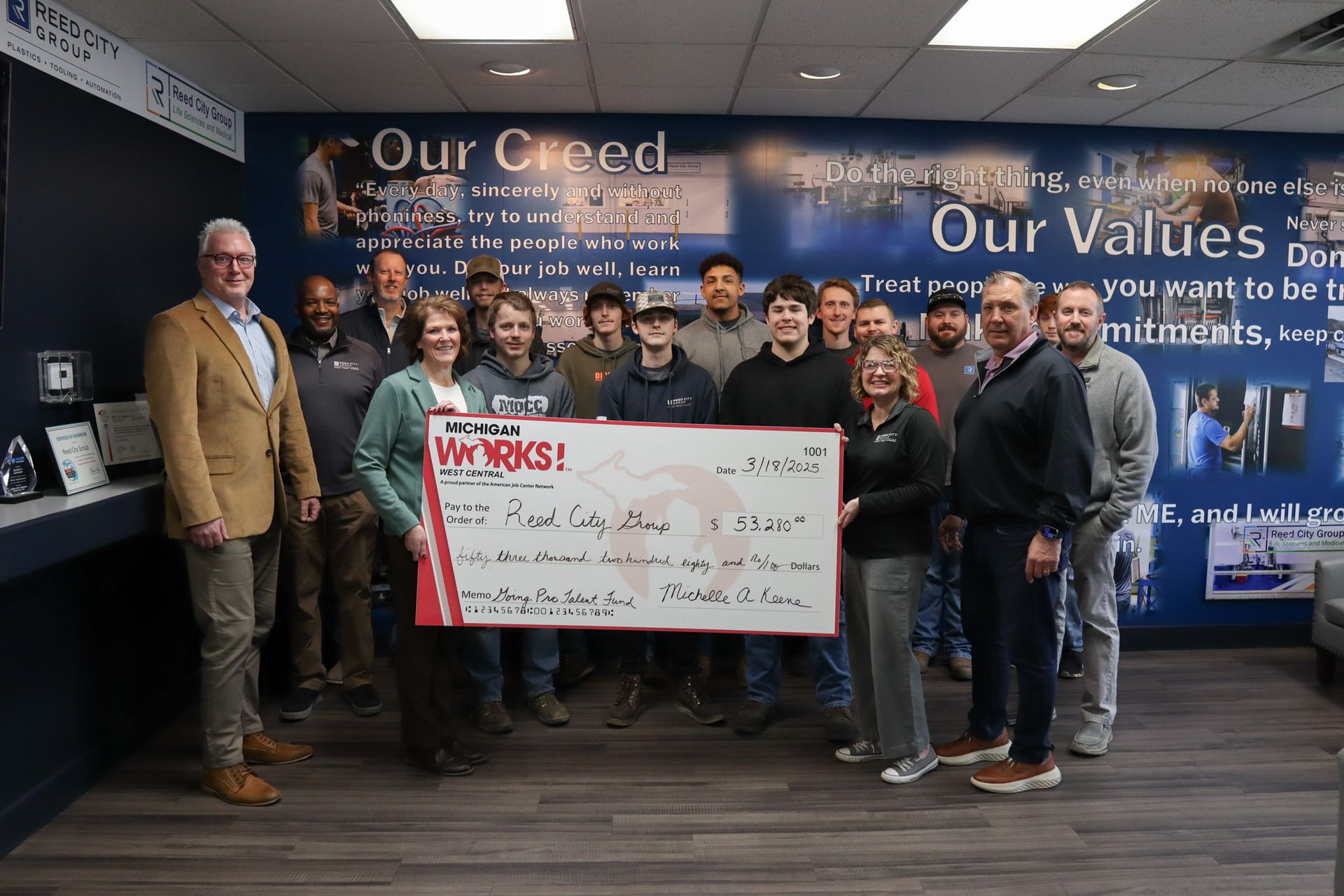 Group of people posing indoors behind a large check in front of a wall reading “Our Creed” and “Our Values”