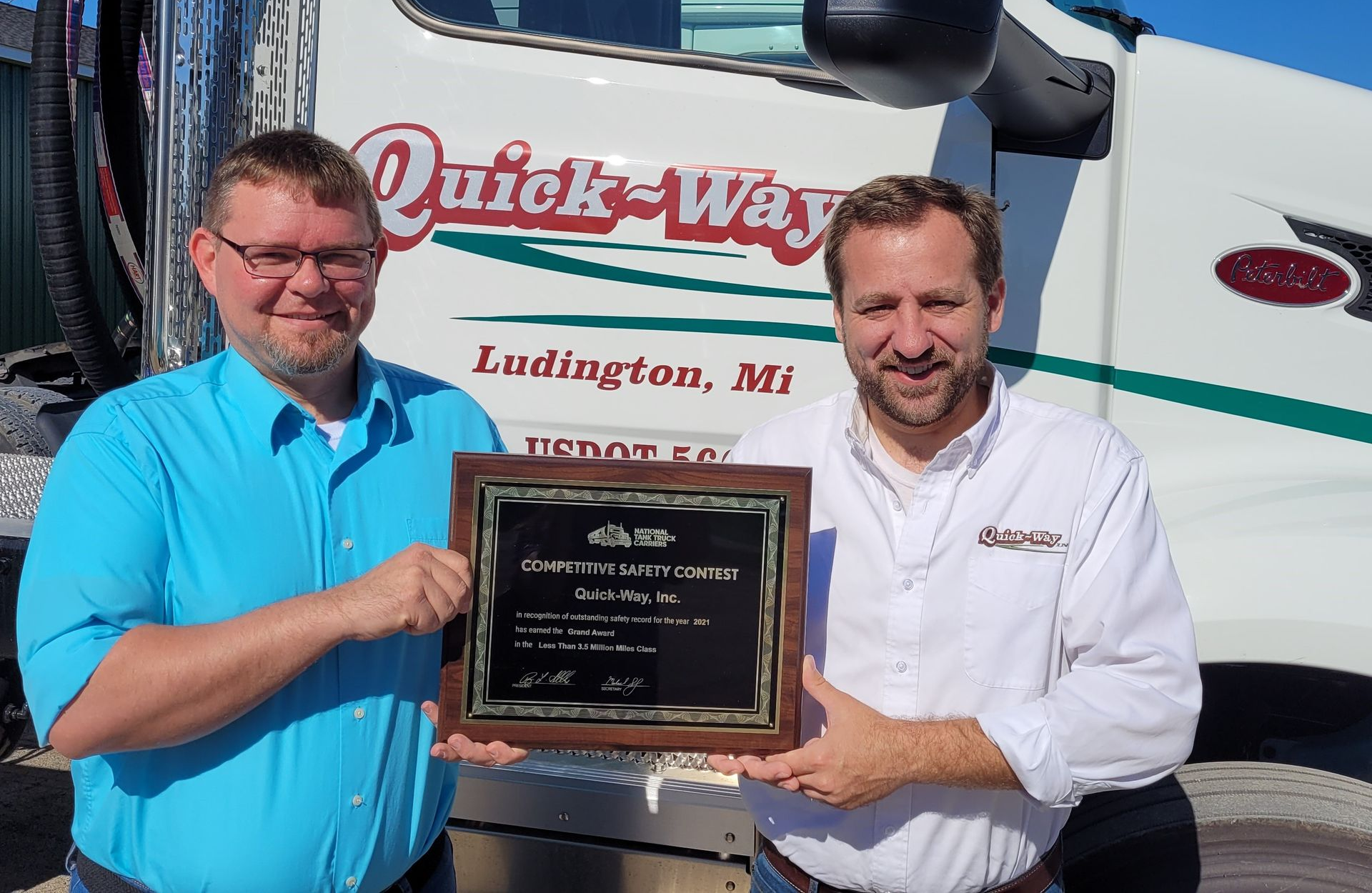 Two men holding a plaque in front of a Quick-Way truck, both smiling.