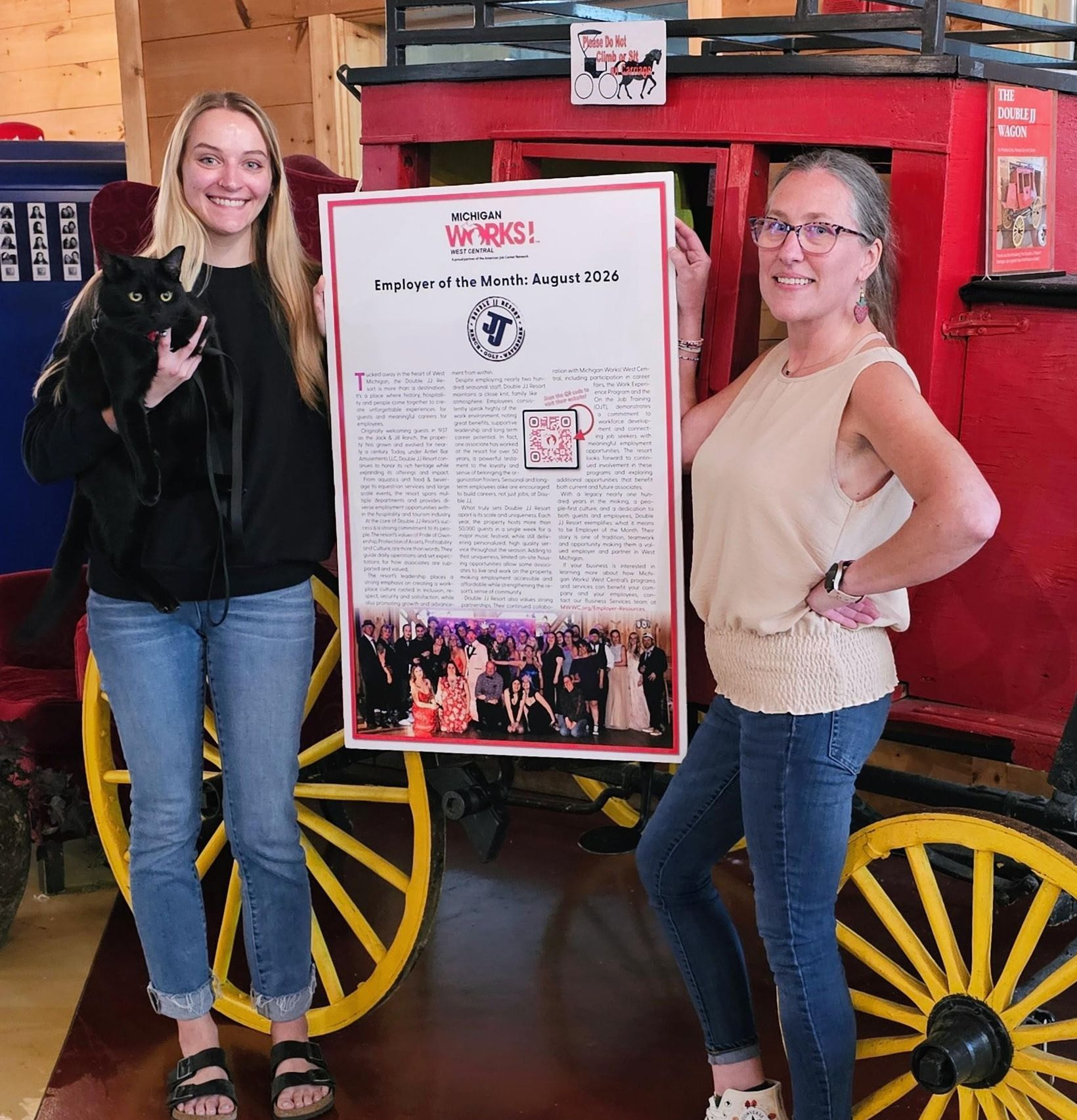 Two women posing beside a framed award poster and a red wagon with yellow wheels in a barn-like setting
