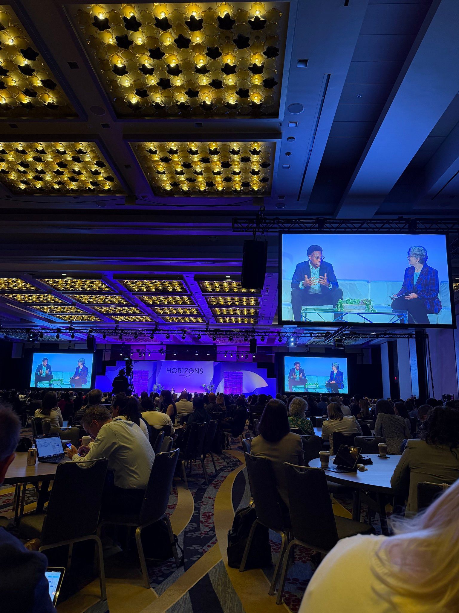 Conference ballroom with stage speakers, blue-purple lighting, and seated audience watching large screens