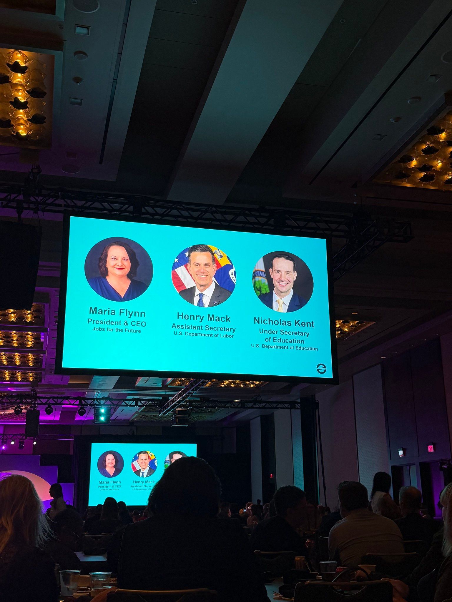 Conference audience facing a large turquoise presentation screen with three speaker headshots.