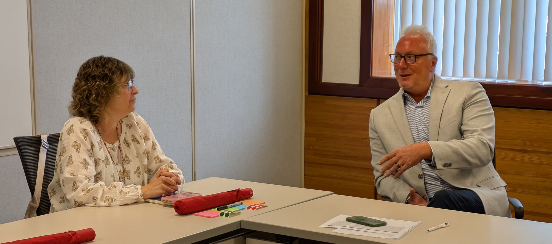 Two people talking across a table in a conference room, with papers and a red folder on the table.