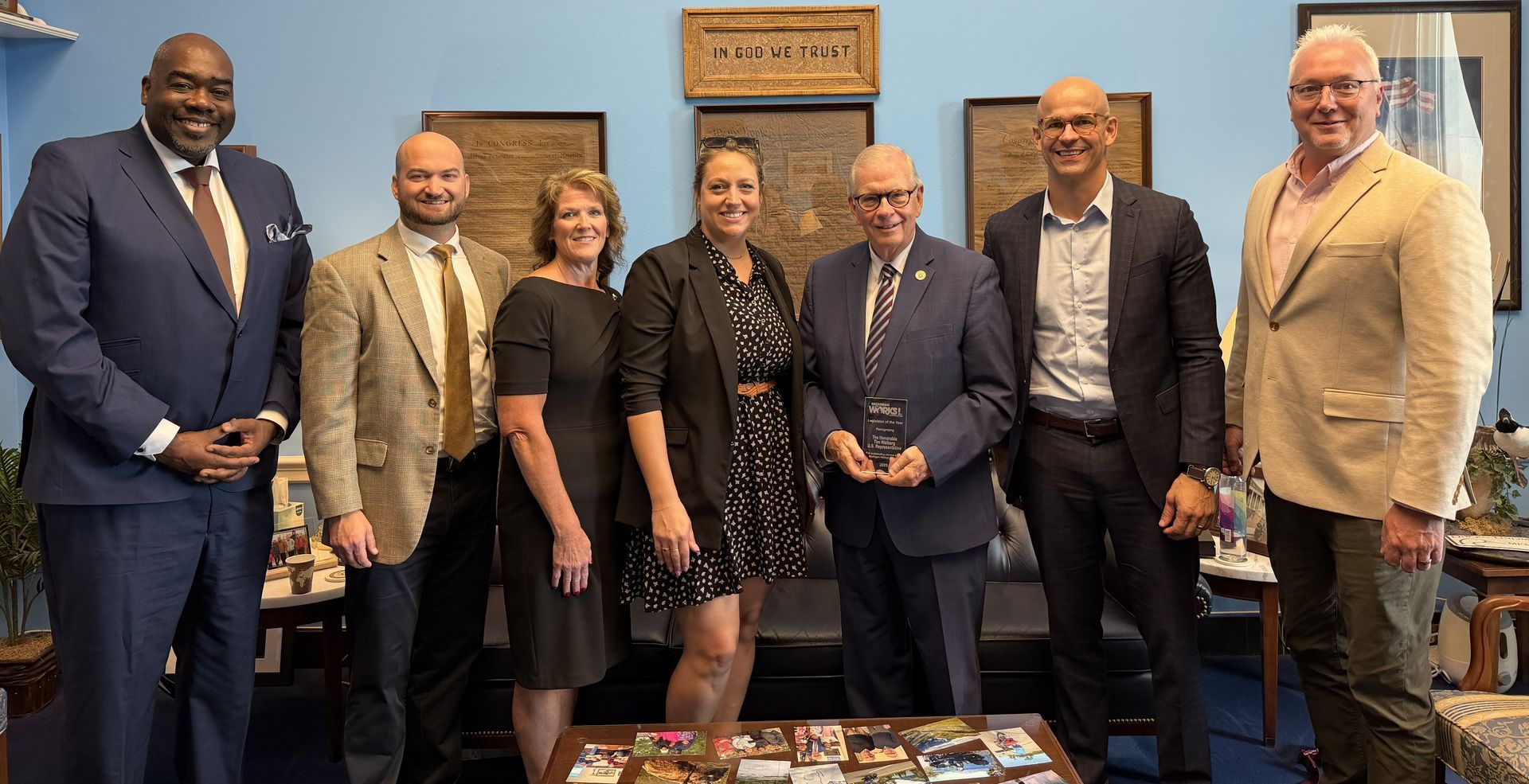 Seven people in business attire posing in an office, smiling beside a desk and framed wall art.