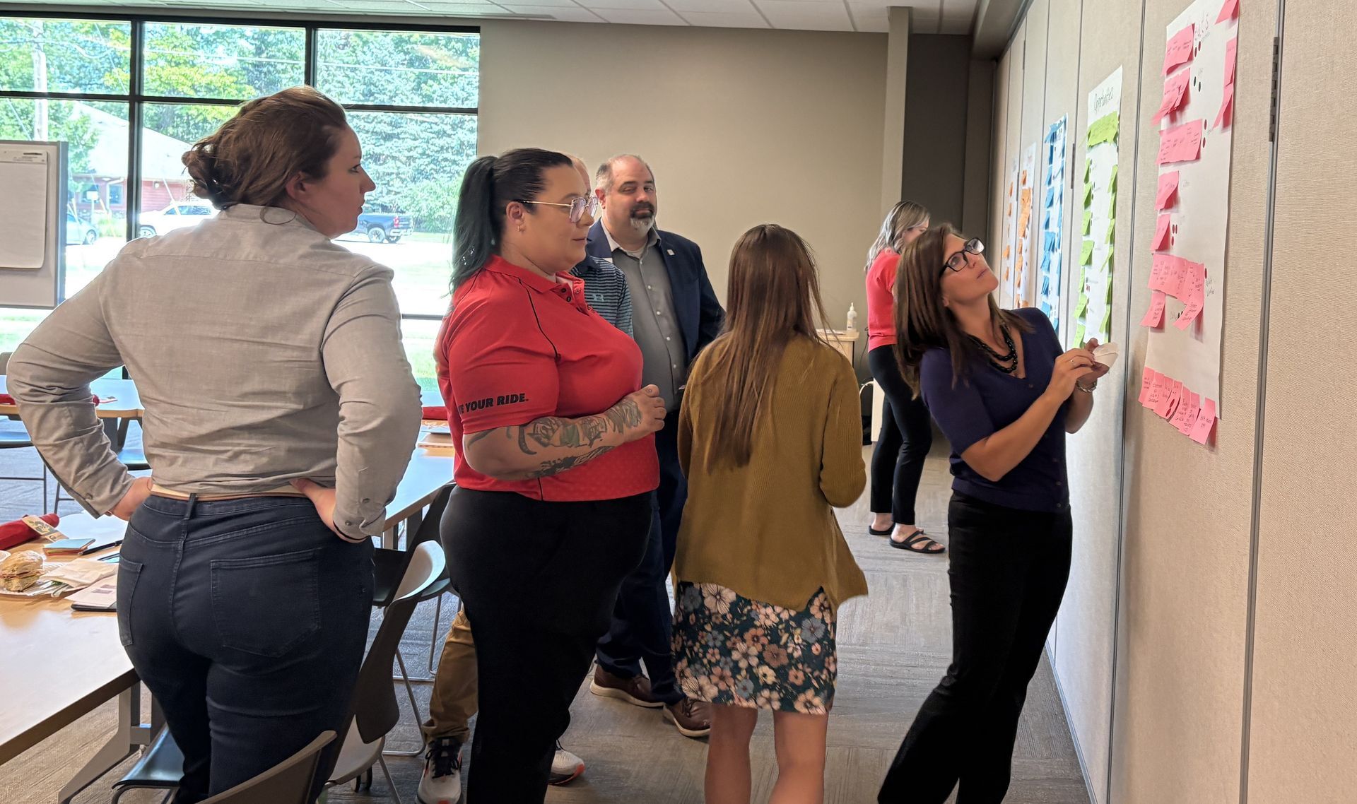 People gathered in a bright room, standing by a wall with pink sticky notes during a collaborative discussion.