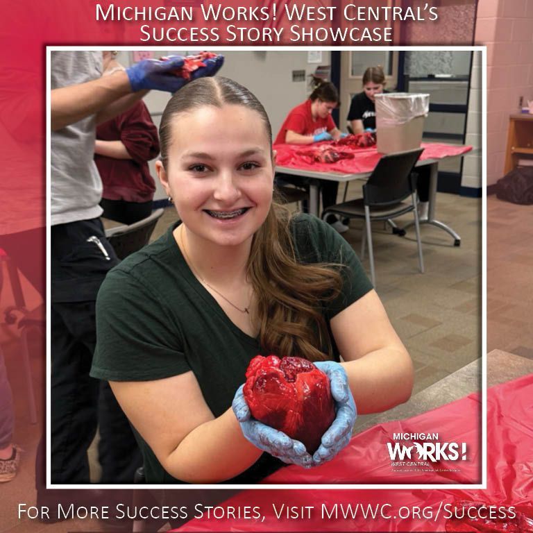 Student smiles while holding a red heart-shaped object in a classroom; Michigan Works! West Central Success Story Showcase.