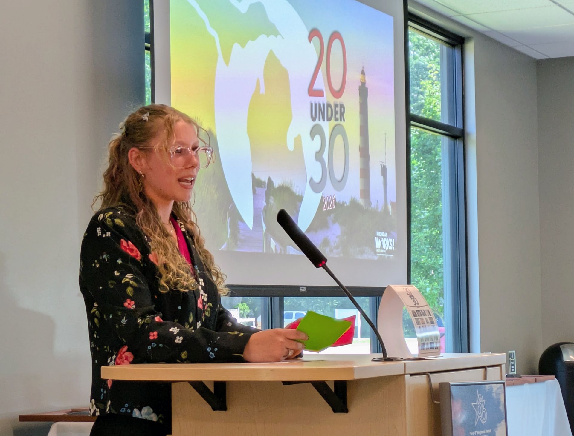 Speaker at podium holding cards, presenting beside a projected “20 under 30” slide in a conference room.