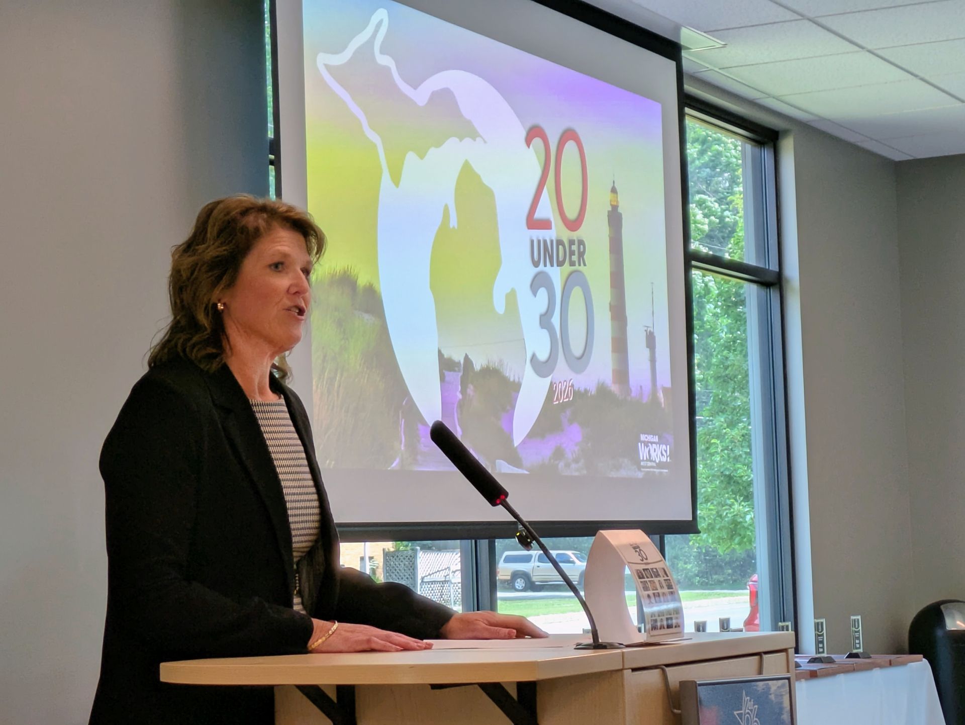 Woman speaking at podium in a conference room, with a projected presentation slide behind her.