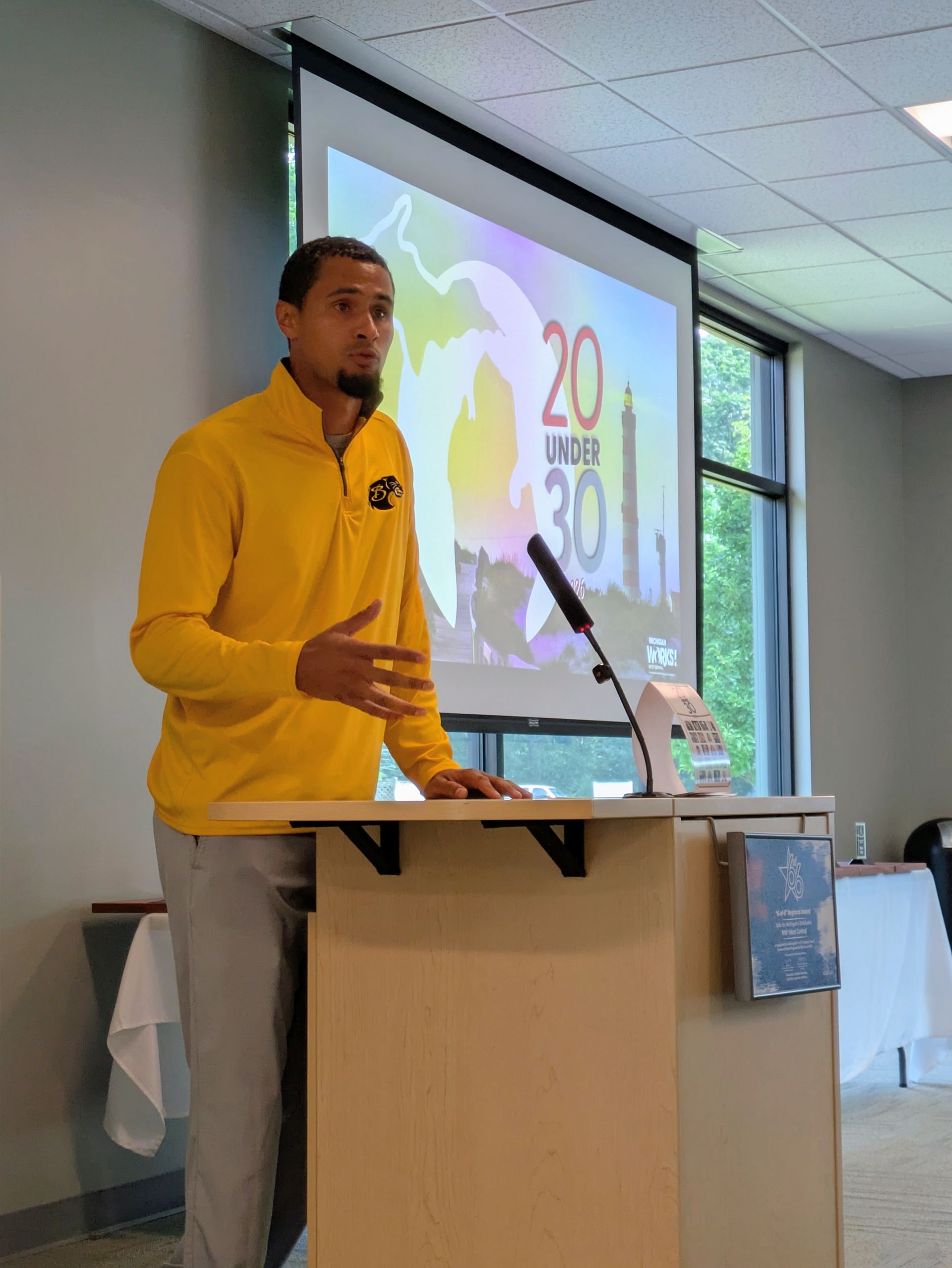 Person in a yellow sweatshirt speaking at a podium during a presentation in a classroom.