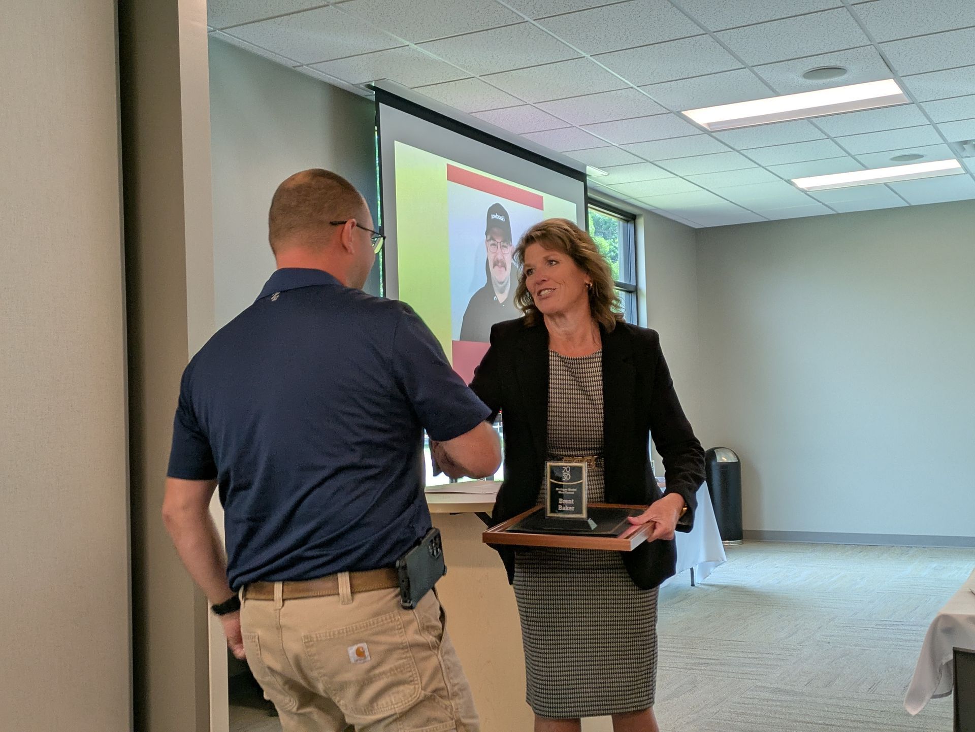 Man handing a plaque to a woman in a hallway, with a presentation screen in the background