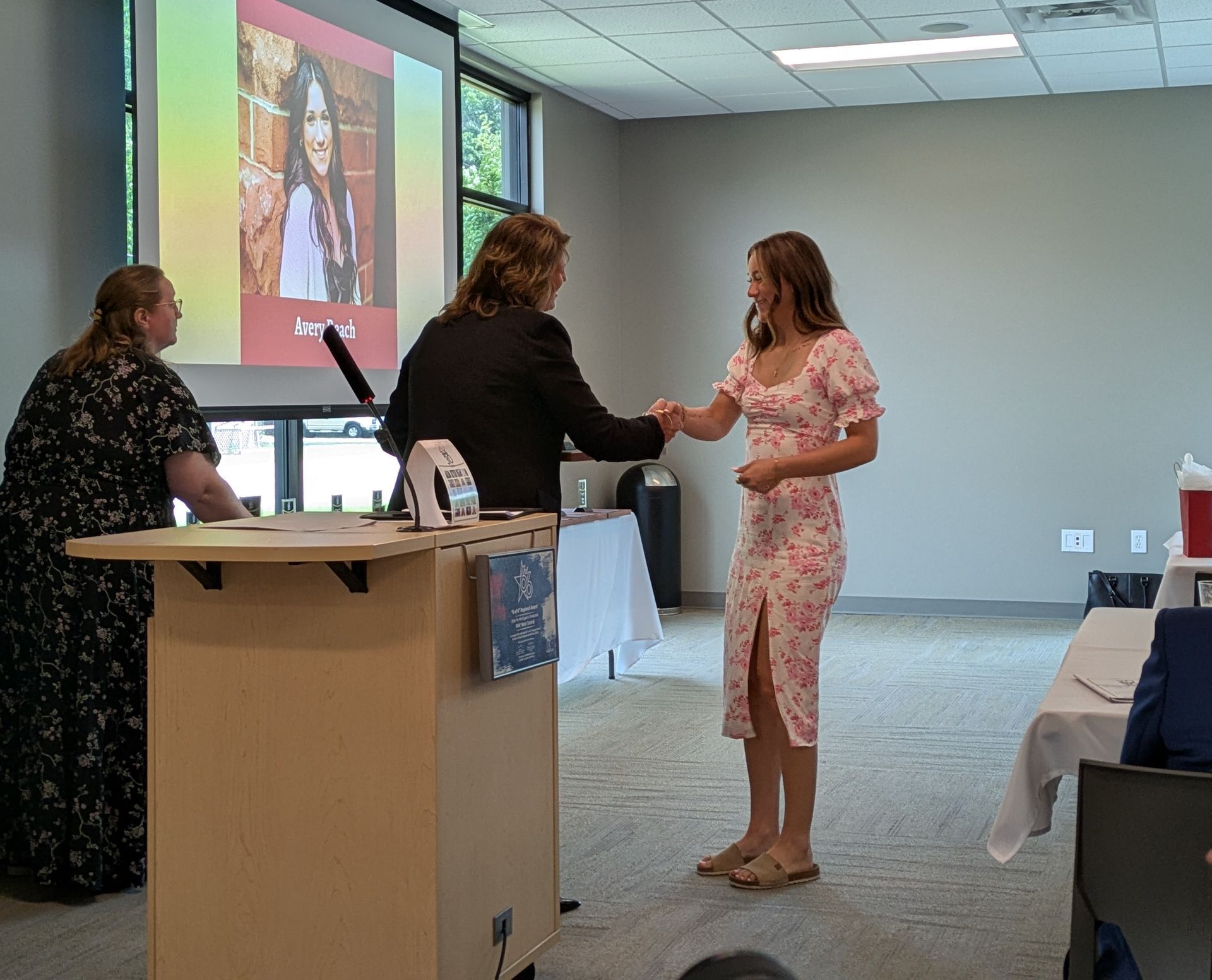 Two women shaking hands at a podium during a presentation in a conference room