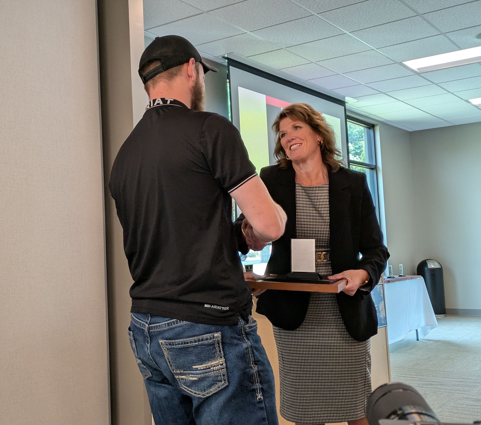 Two people exchanging a tray in a bright meeting room, one smiling by a projection screen.
