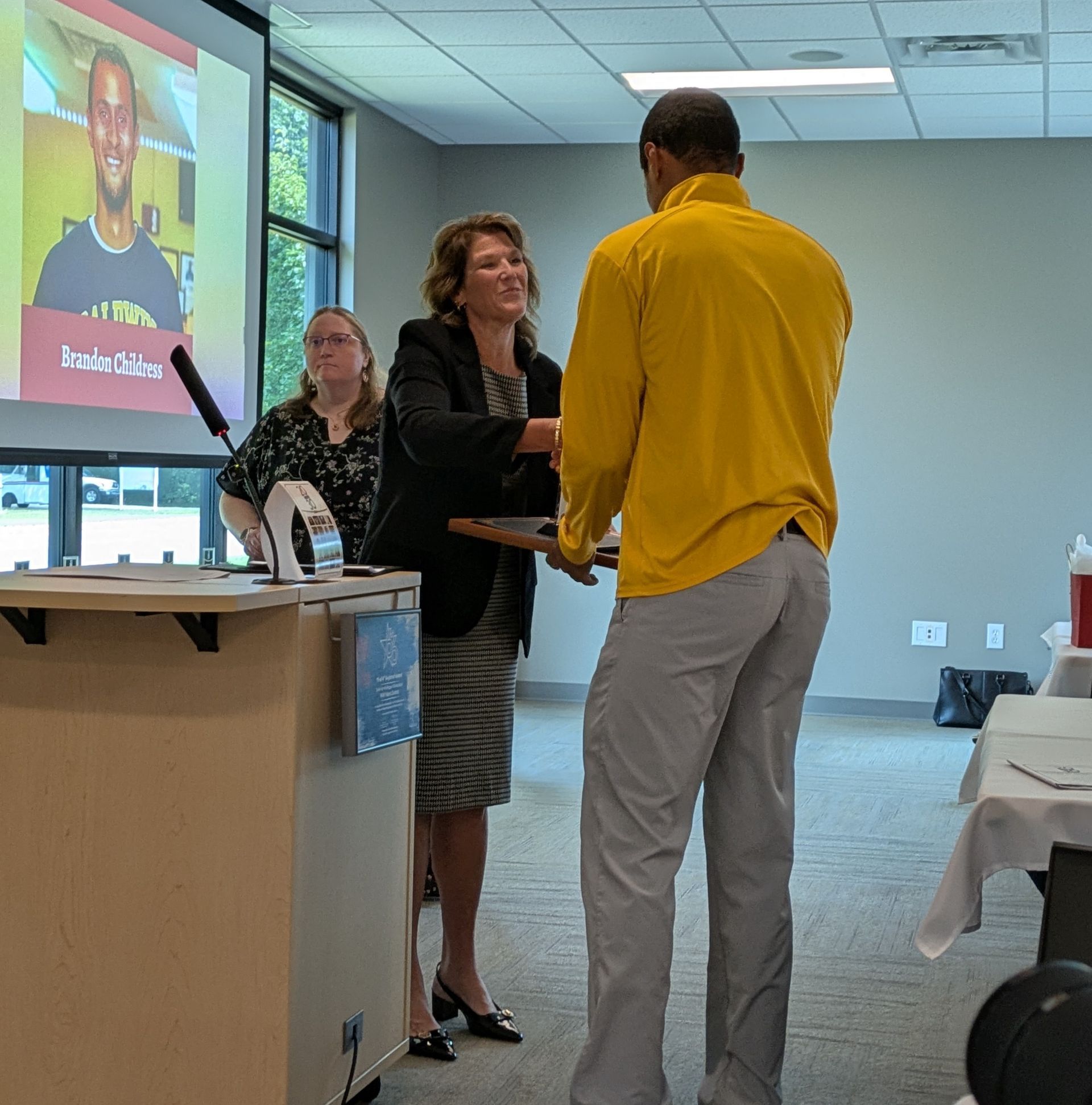 Two people shake hands at a podium in a conference room with a projected presentation screen.