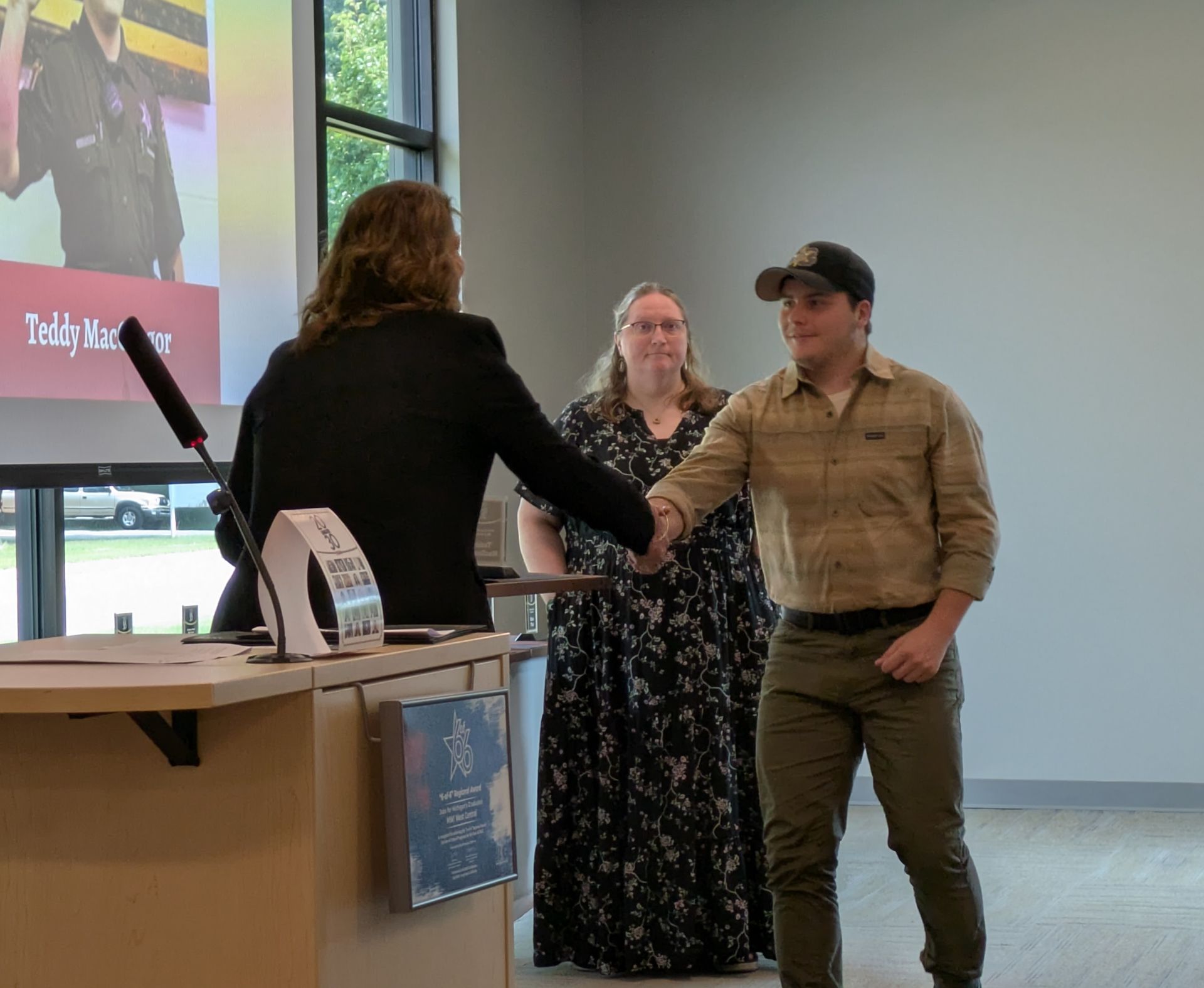 Two people shaking hands at a counter in a bright indoor lobby, with a presentation screen in the background.
