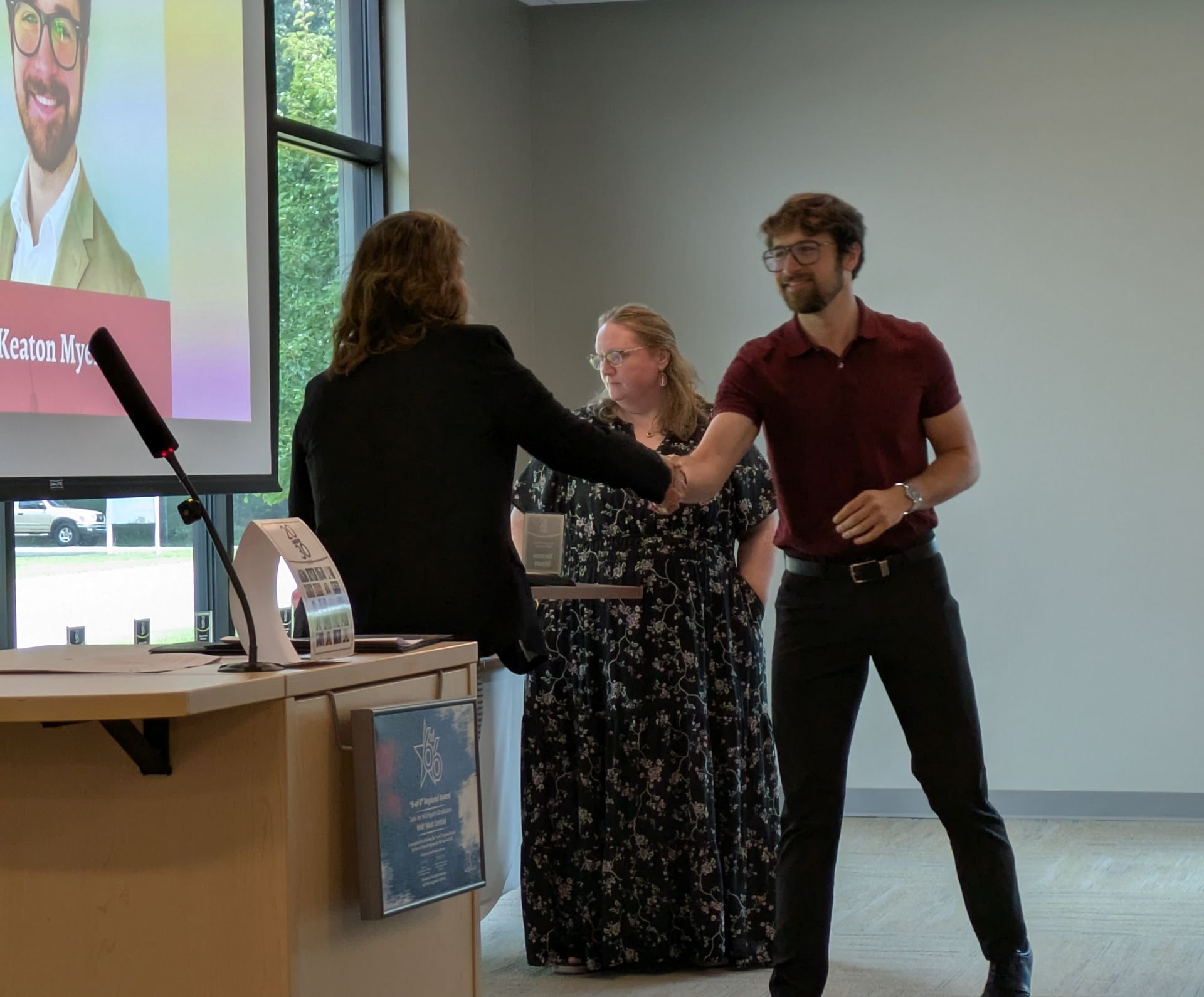 Two people shaking hands at a podium during an indoor presentation, with a projected slide in the background.