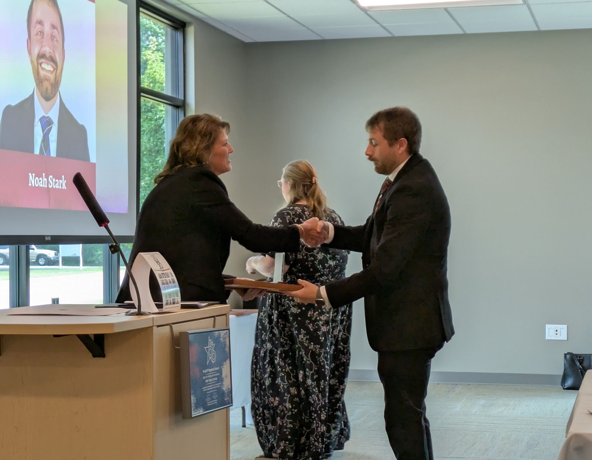 Two people shaking hands at a conference table in a bright room with a presentation screen nearby