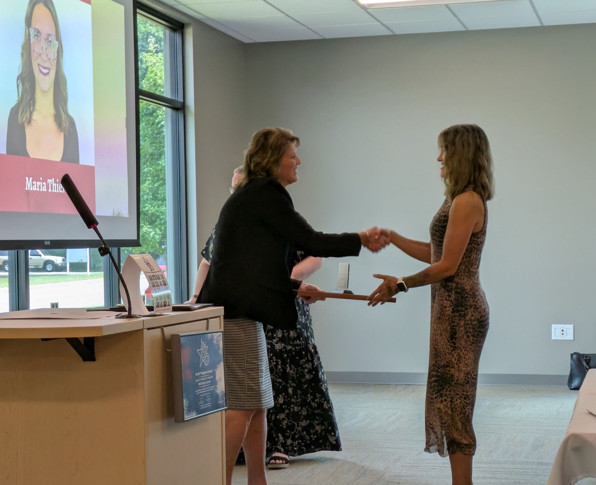 Two people shake hands at an indoor ceremony beside a projected speaker screen.