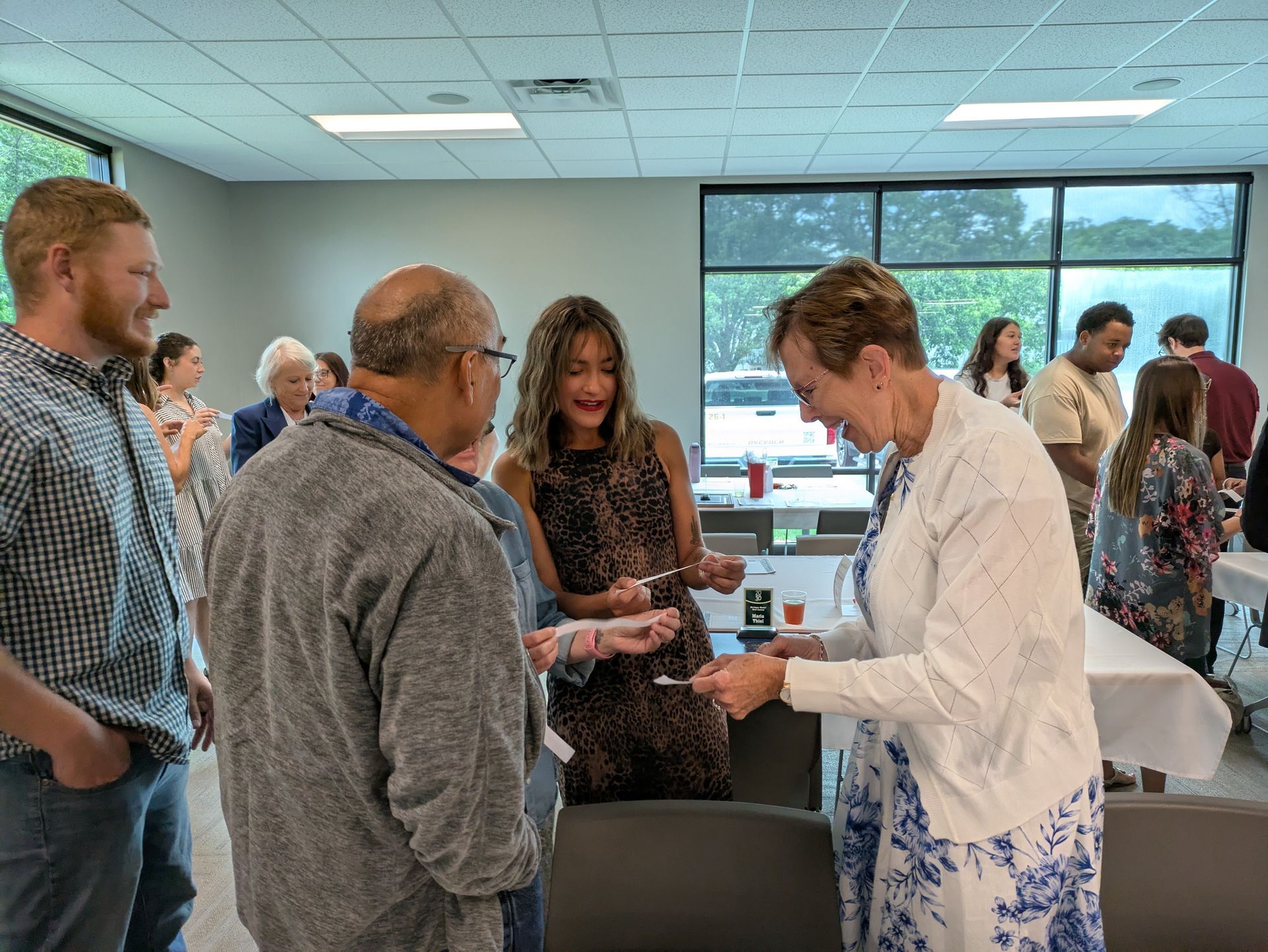 People gathered in a bright room, talking and examining papers at a social event.