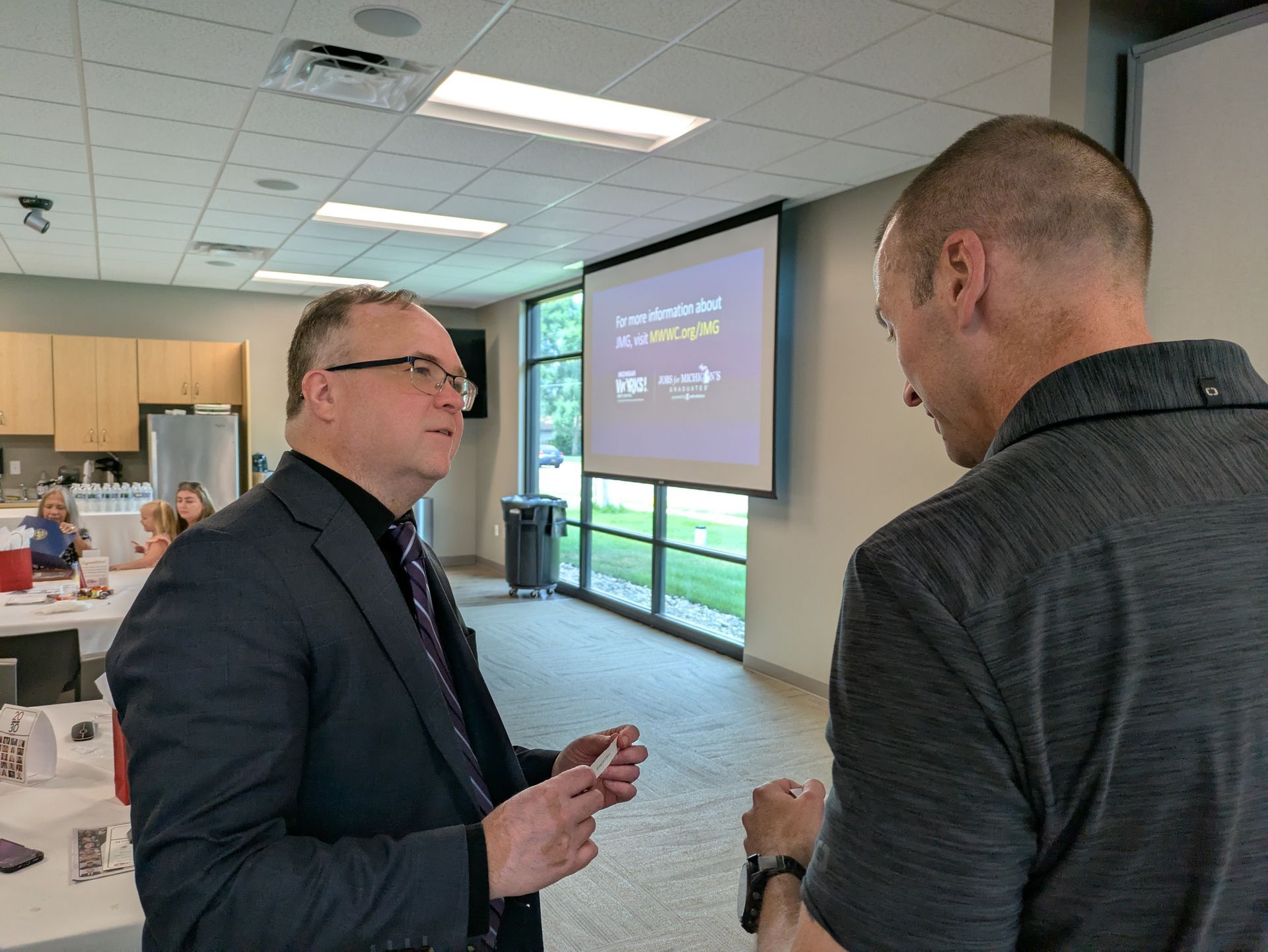 Two men talking in a bright conference room near a presentation screen