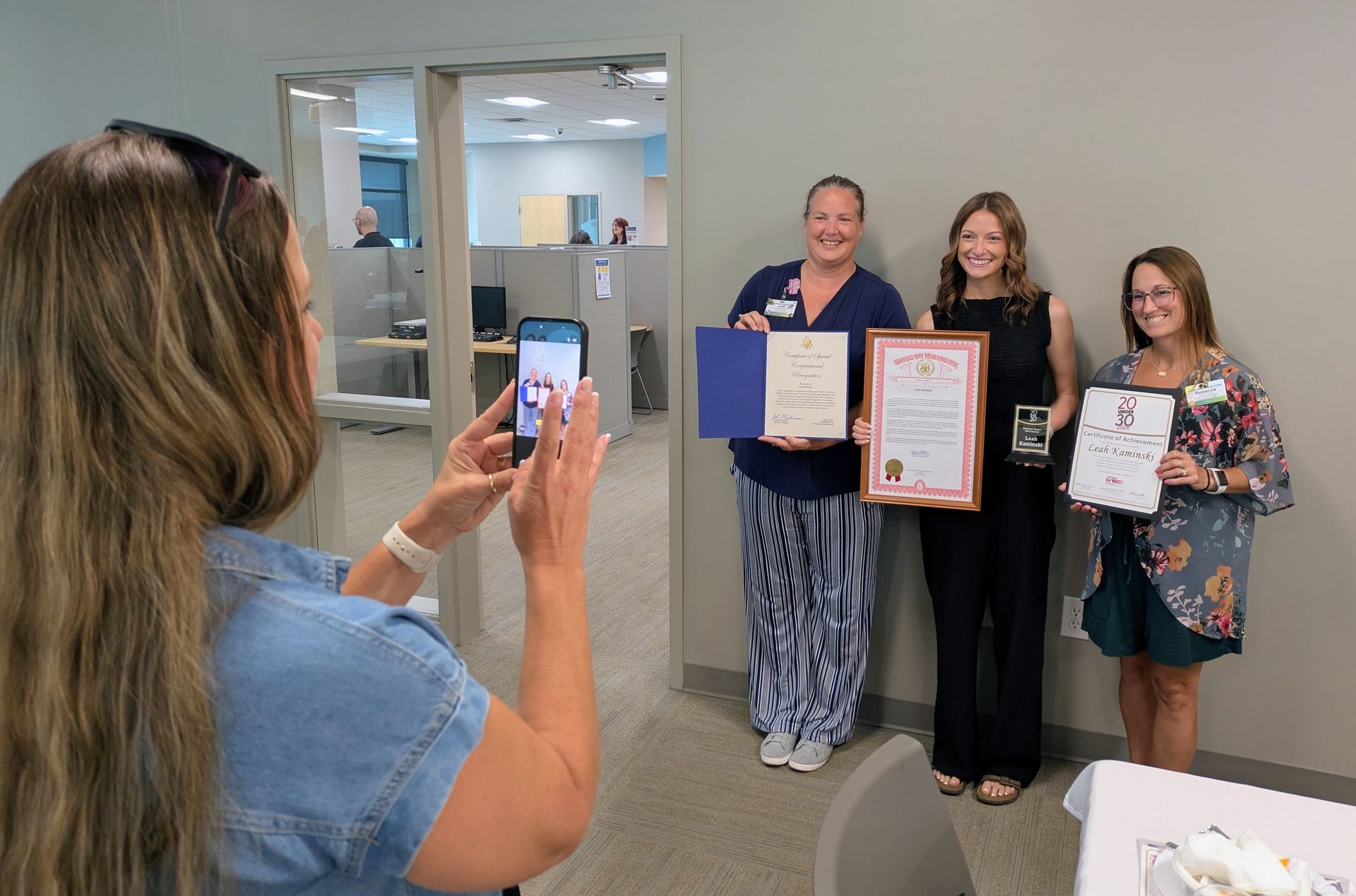 Woman takes a photo of three people posing with certificates in an office hallway.