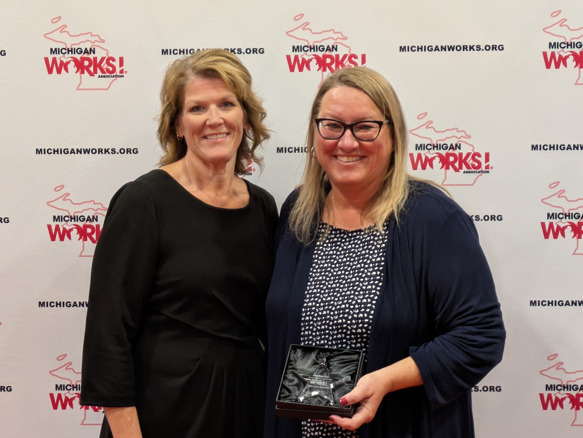 Two women posing at a McDonald’s Works event, one holding a plaque, in front of a logo backdrop