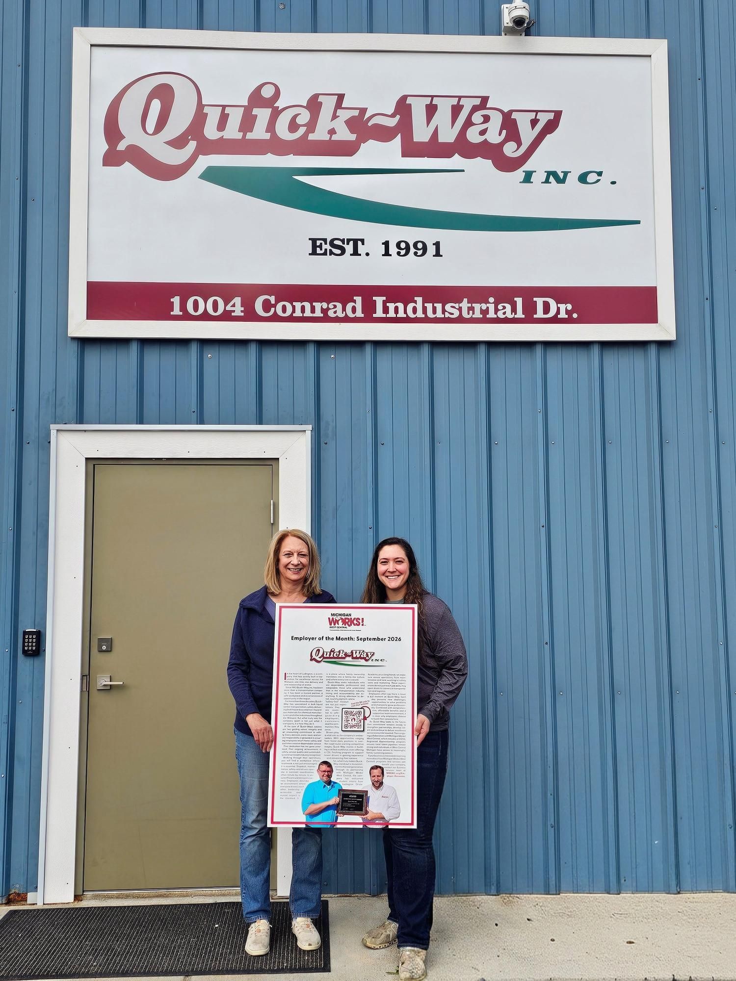 Two women holding a poster in front of a Quick-Way Inc. sign on a blue building