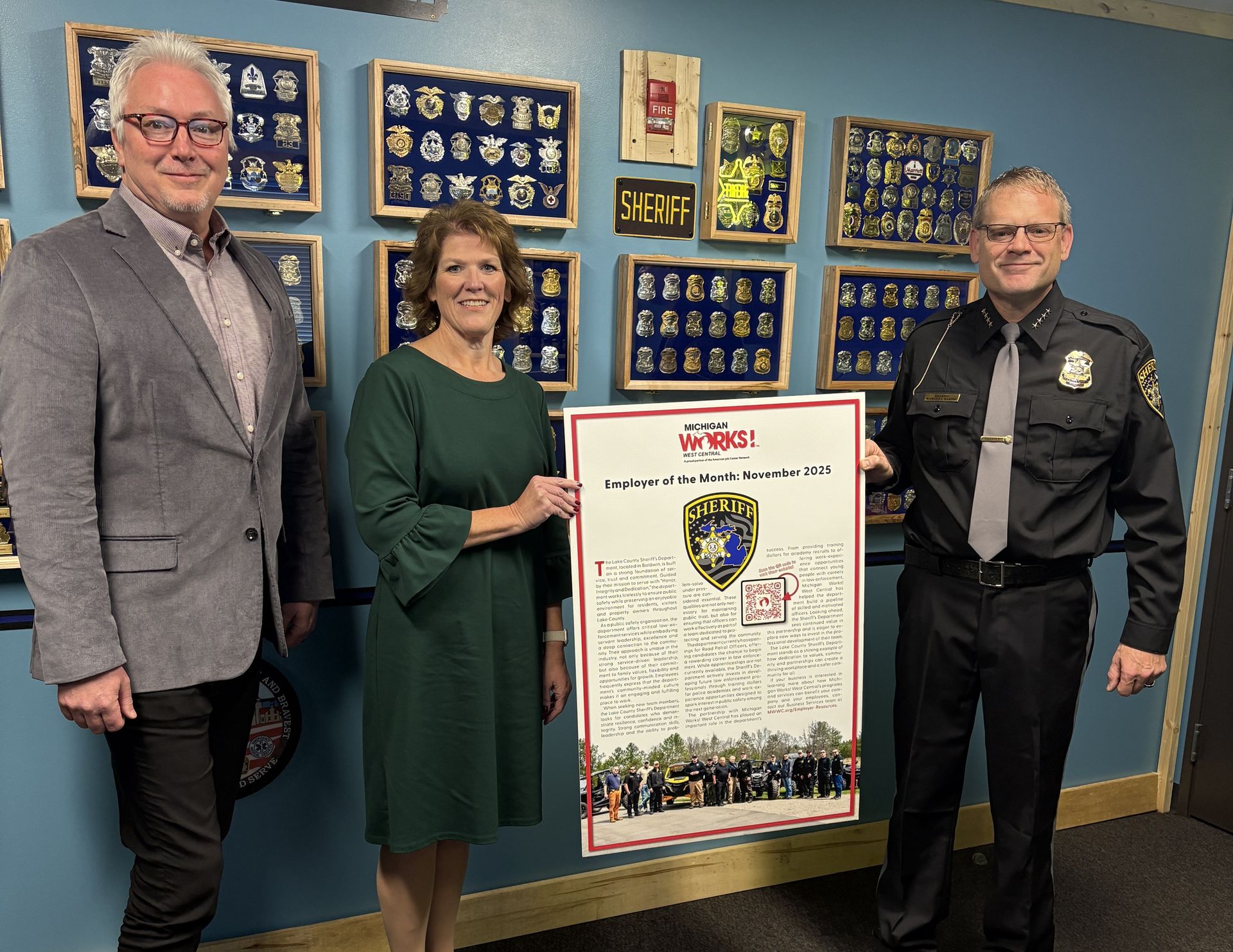 Three people pose indoors beside a framed certificate and awards wall.