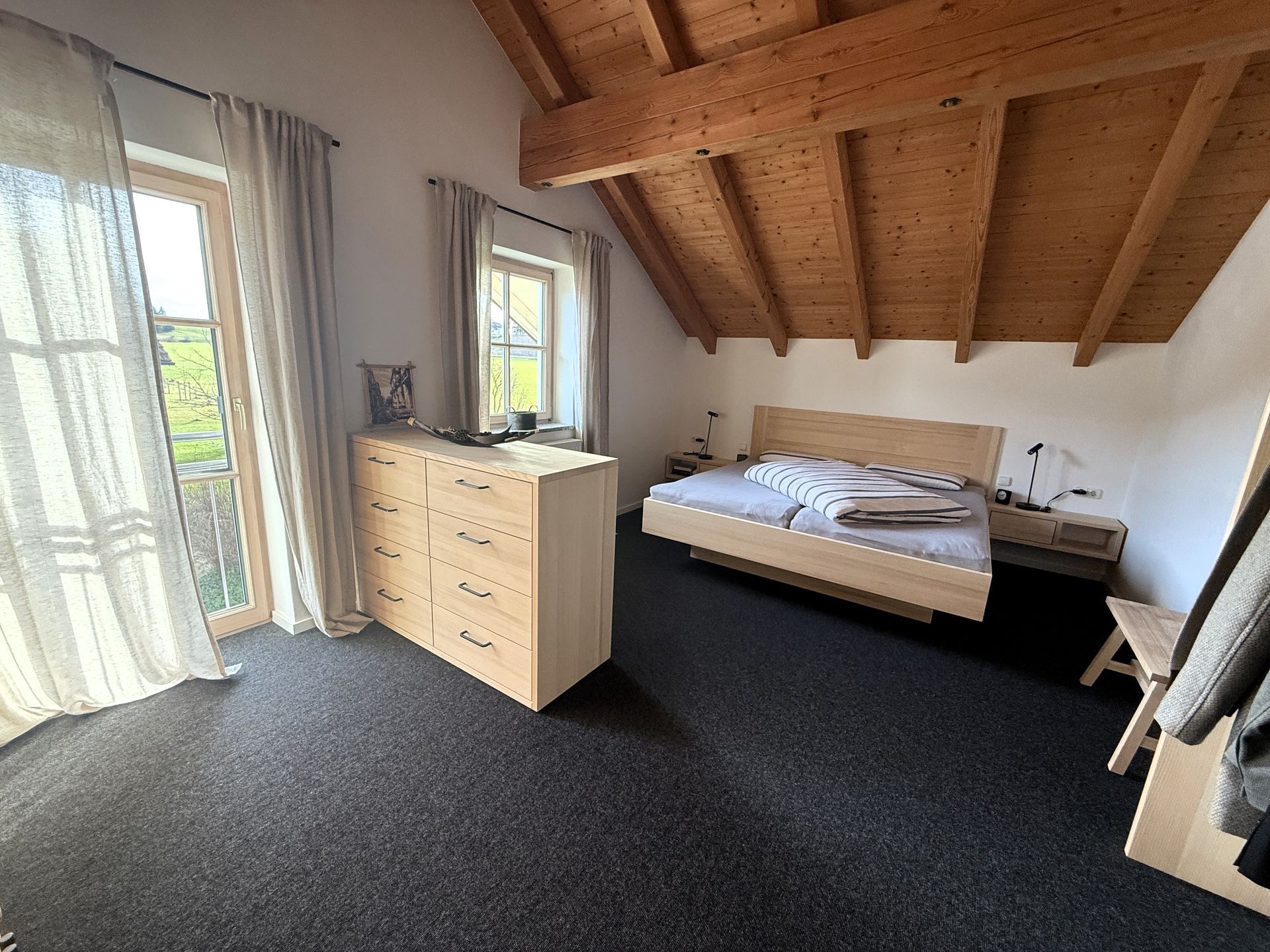Helles Schlafzimmer mit schräger Holzdecke, Bett, Kommode und großen Fenstern mit Vorhängen