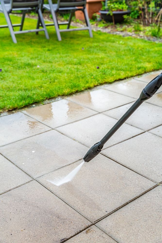 Pressure washing concrete patio, jet of water spraying tiles, near green lawn and chairs.