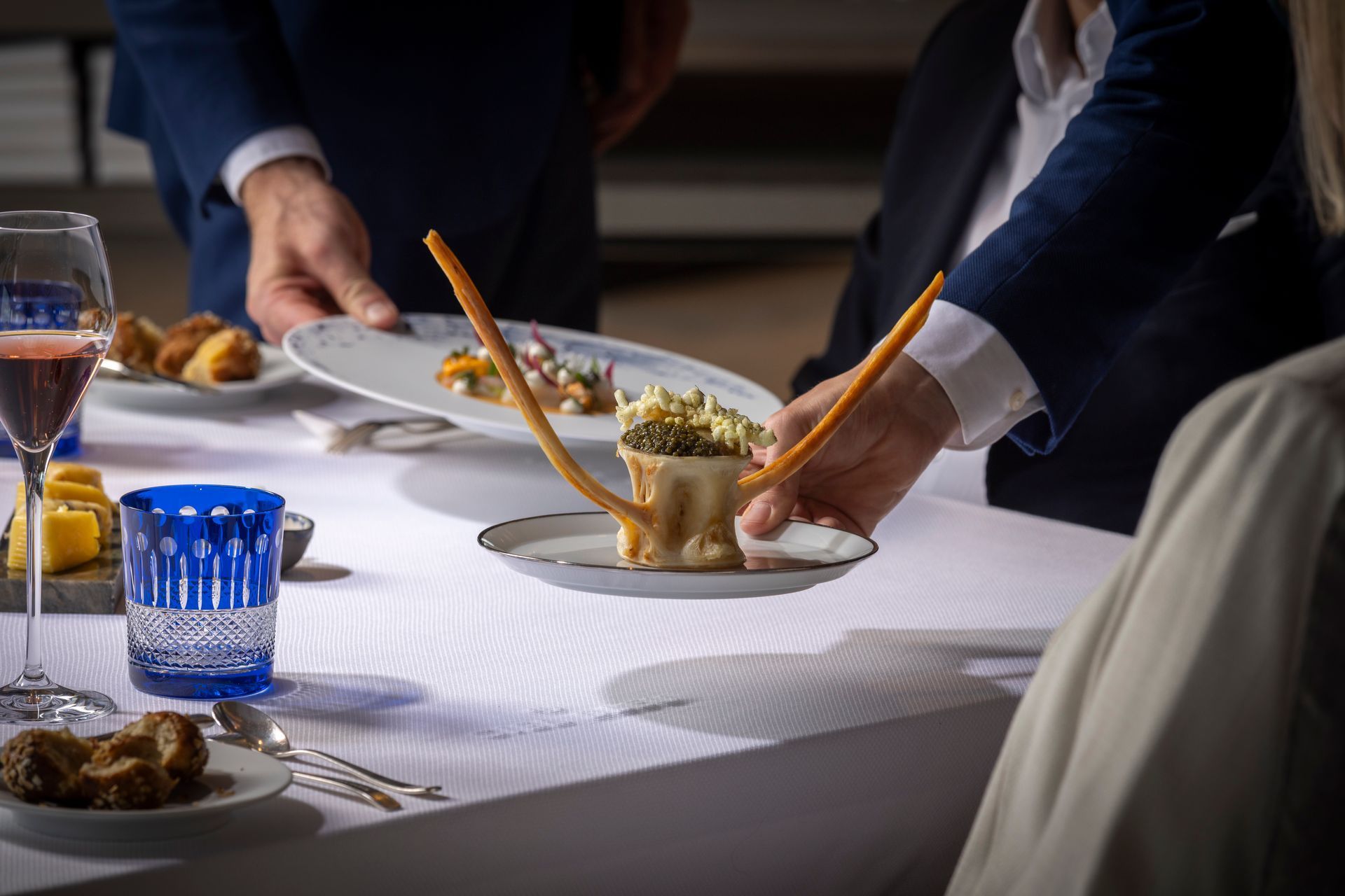 Hands serving plated food at a formal dining table with white tablecloth and blue glasses