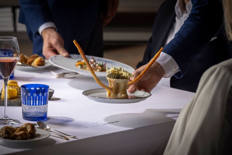 Hands serving plated food at a formal dining table with white tablecloth and blue glasses