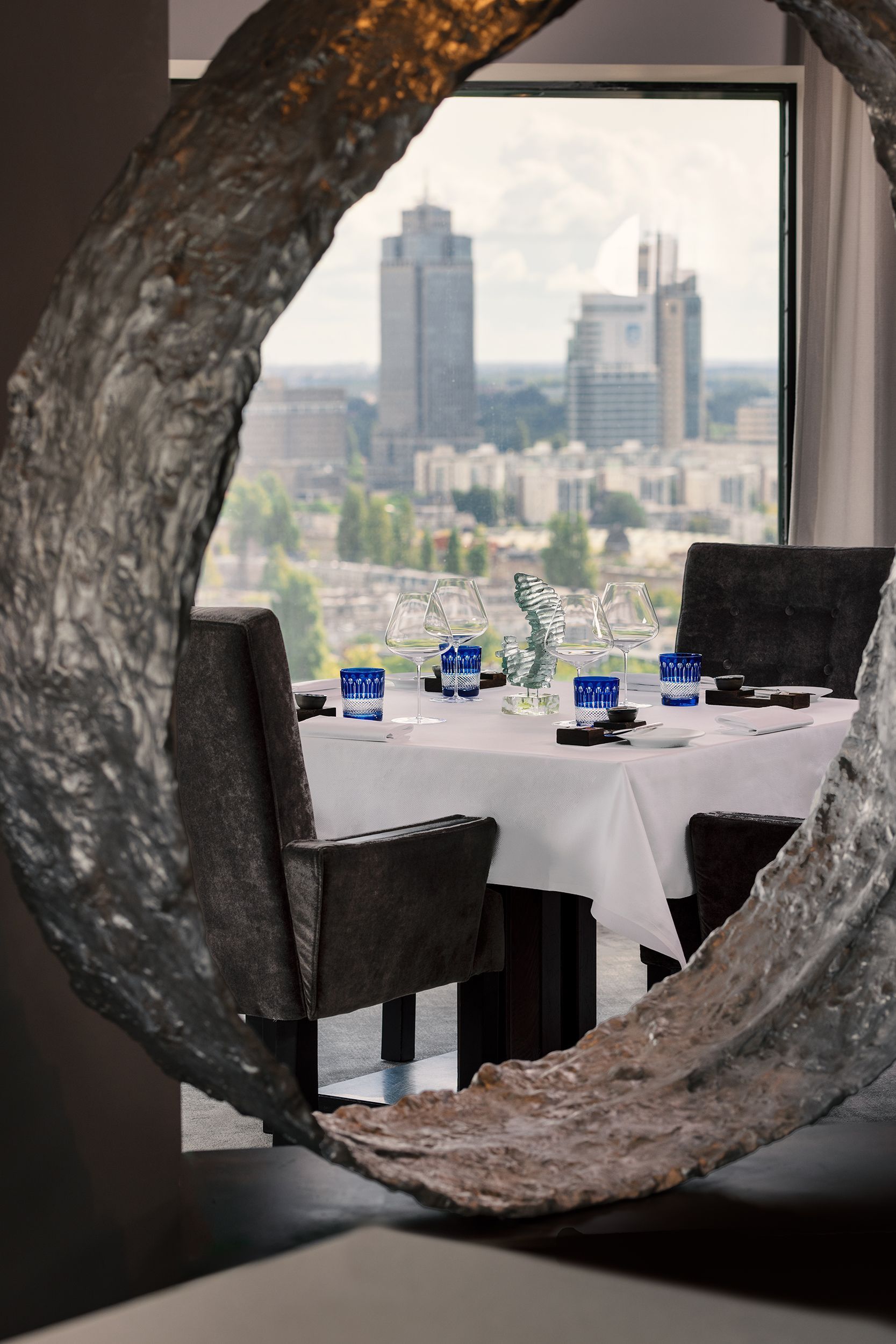 Restaurant table framed by a circular stone opening, with city skyline visible beyond the window.