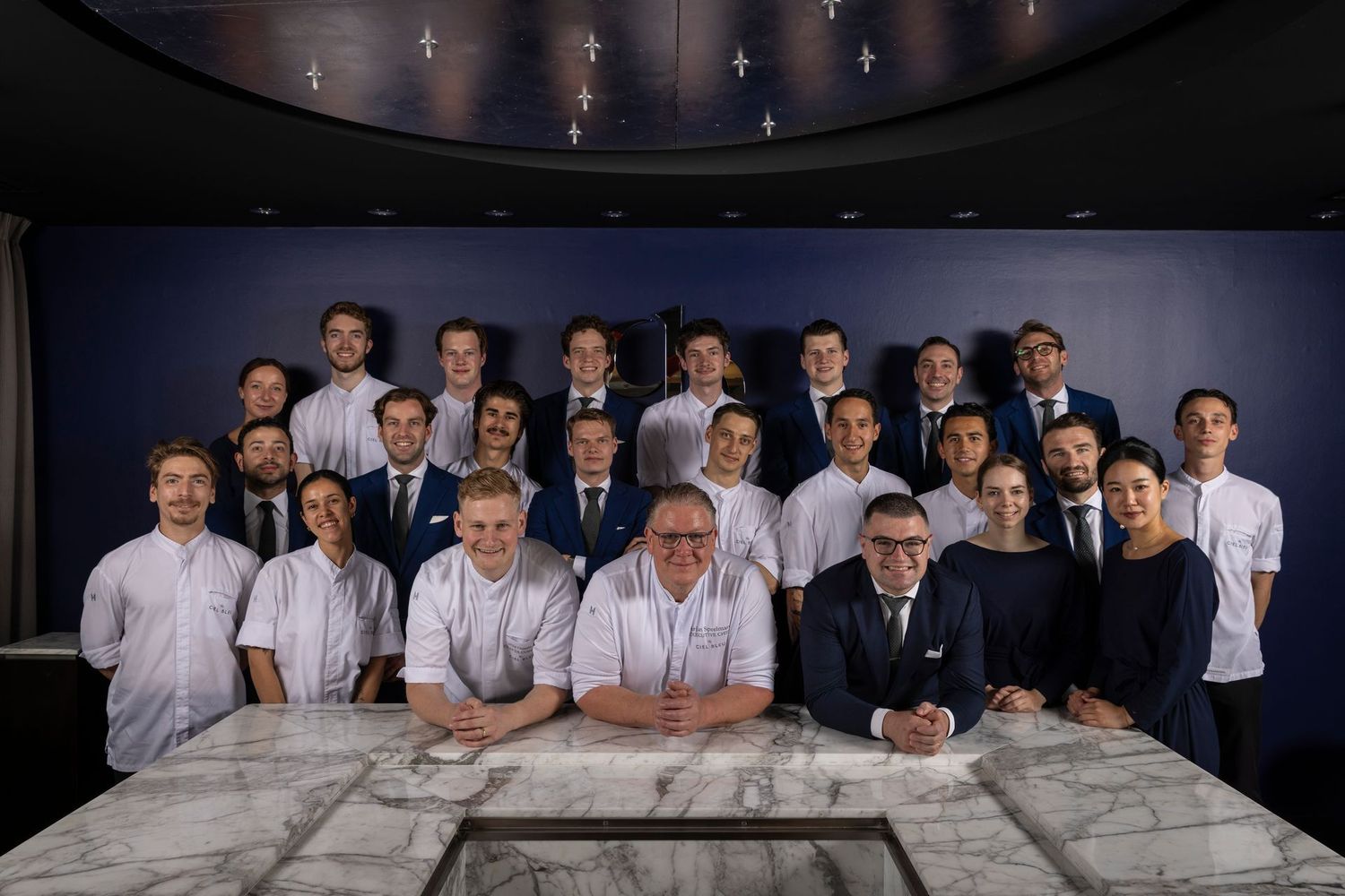 Large group of professionals posing in a conference room behind a marble table, with a dark ceiling and wall backdrop