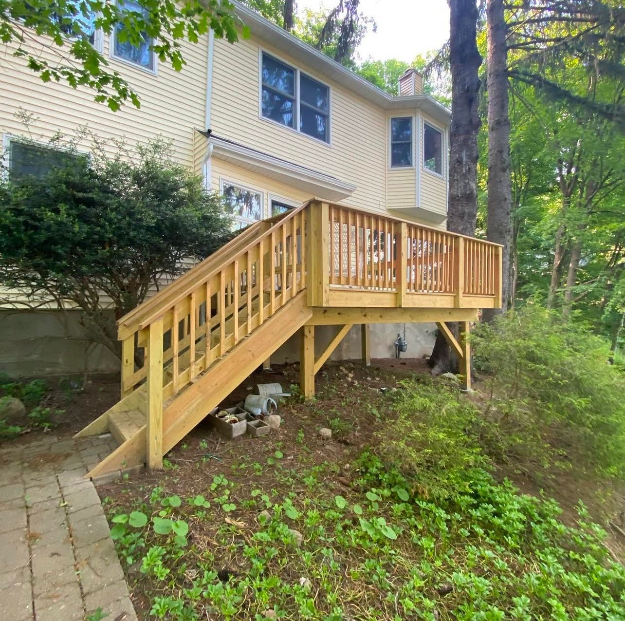 Wooden deck and stairs attached to a light yellow two-story house, surrounded by greenery.