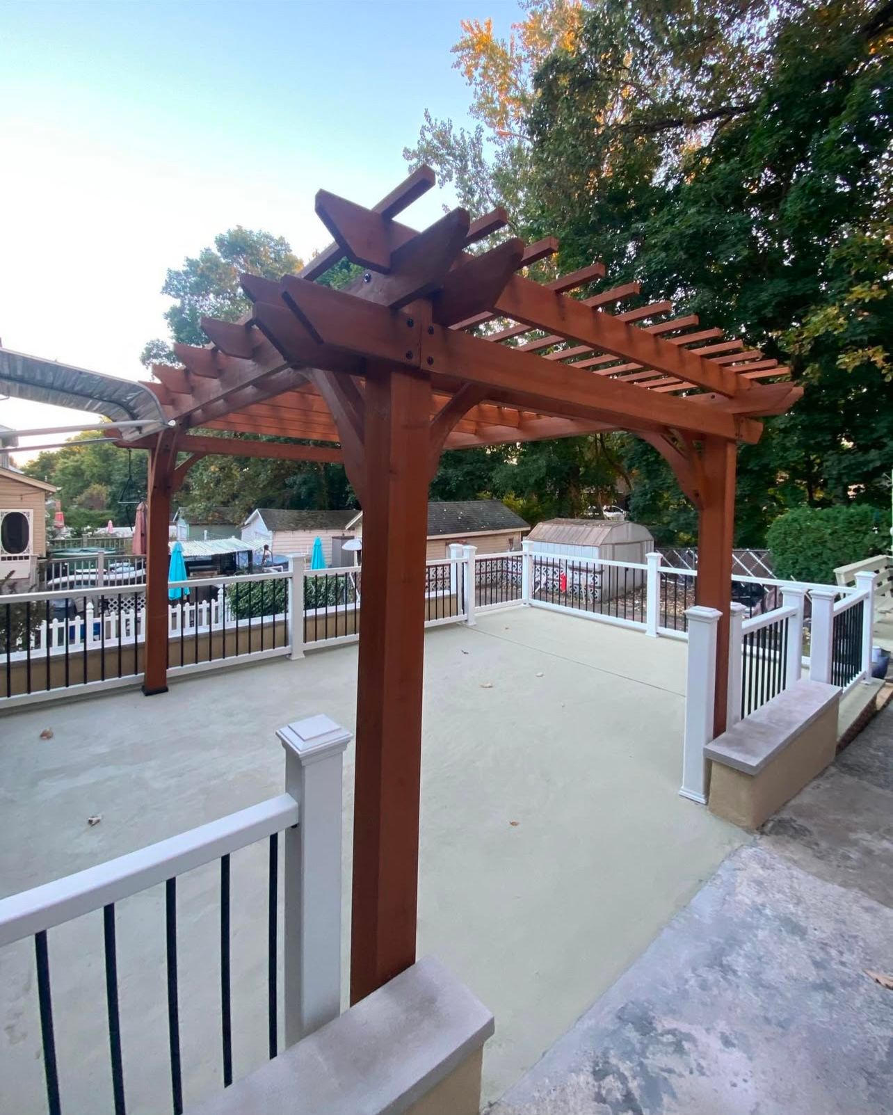 Wooden pergola on a deck, painted brown with white railings. Green trees in the background.