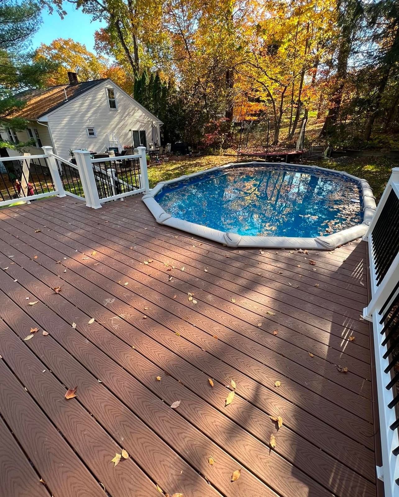 Wooden deck with an above-ground pool in a backyard. Leaves scattered on the deck, surrounded by trees and a house.