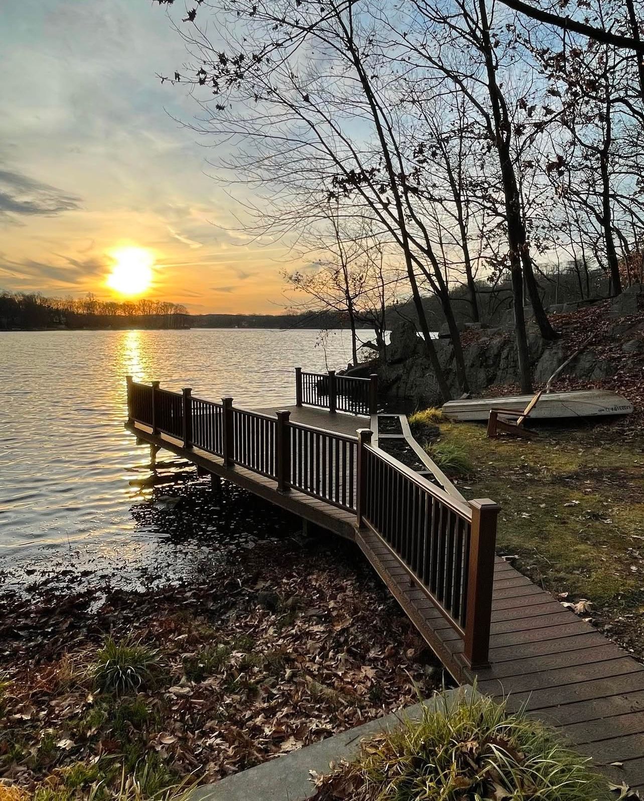 Wooden dock extending into a lake at sunset. Trees line the shore; golden sunlight reflecting on the water.