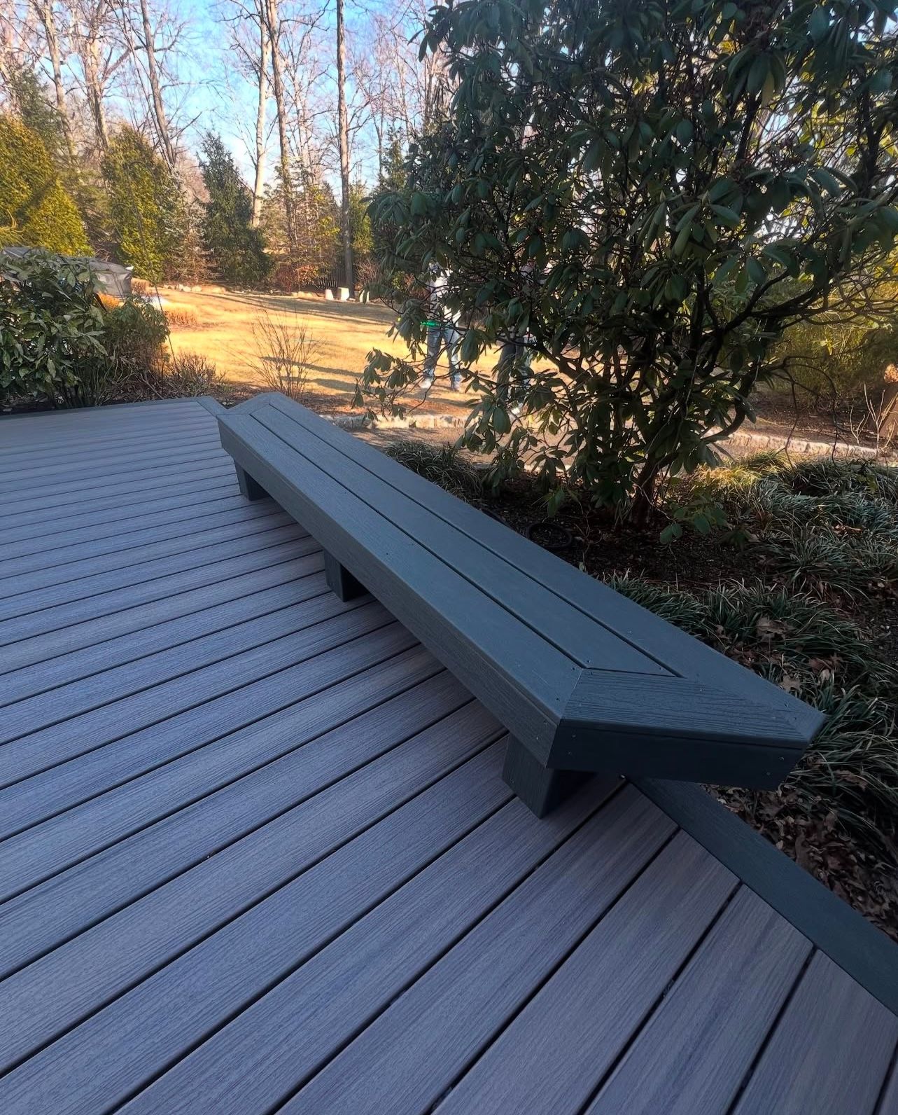 A gray bench on a gray deck, with greenery and a grassy yard in the background on a sunny day.