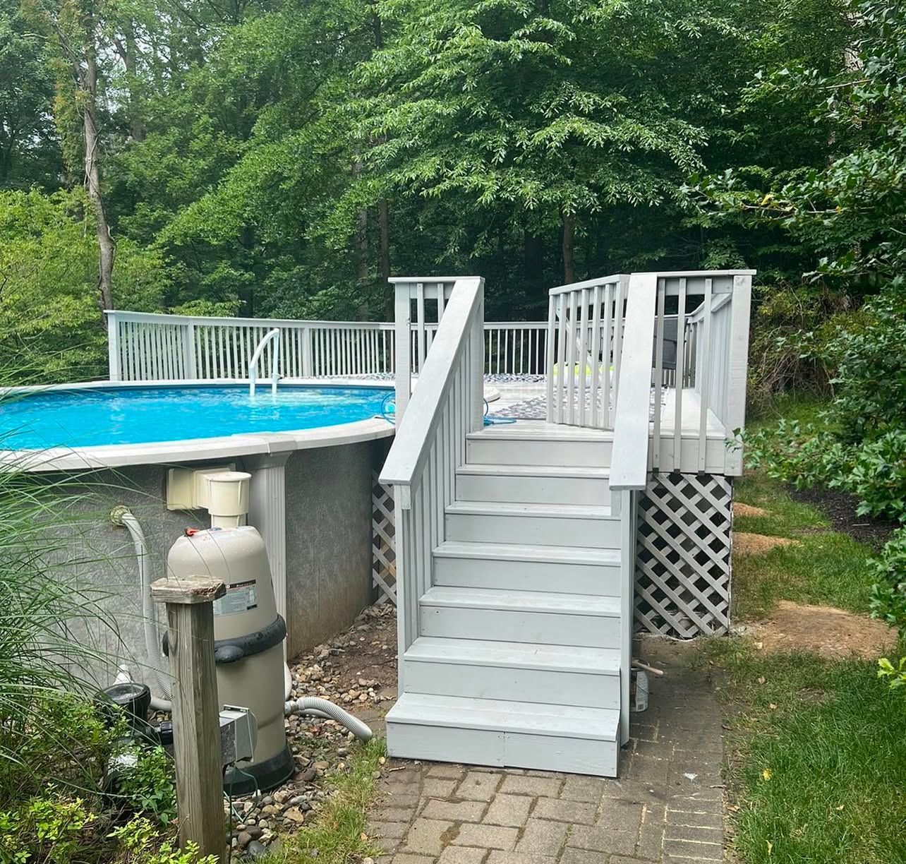 Above-ground pool with gray deck and stairs; surrounded by lush greenery.