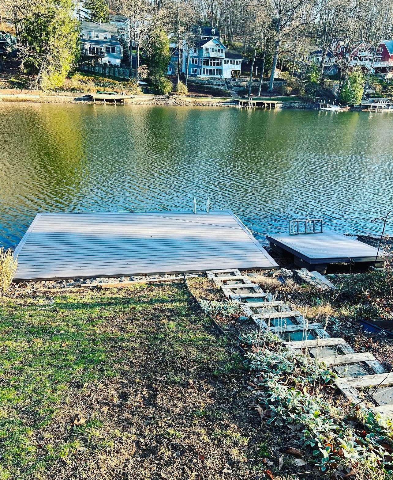 A wooden dock extends into a rippled lake with houses on the opposite shore. A set of steps leads down to the dock.