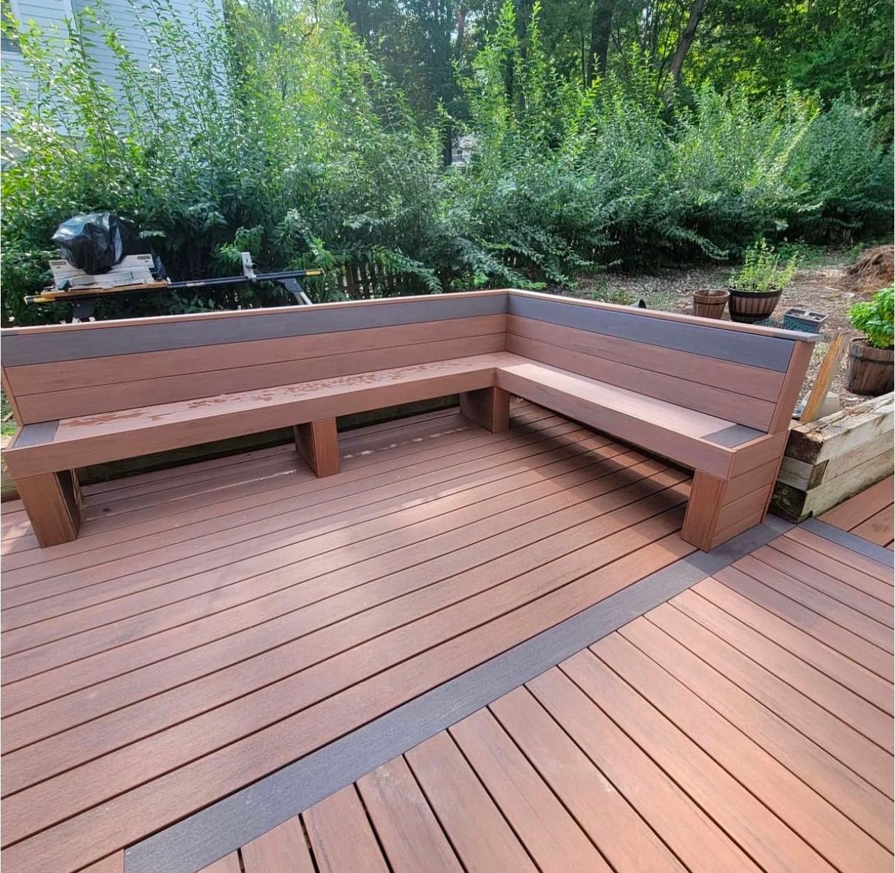 L-shaped wooden outdoor bench on a deck, with brown and gray planks and a green natural background.