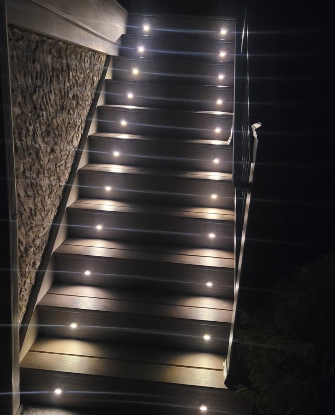 Staircase illuminated by evenly spaced small lights on each step; dark background, stone wall on one side.